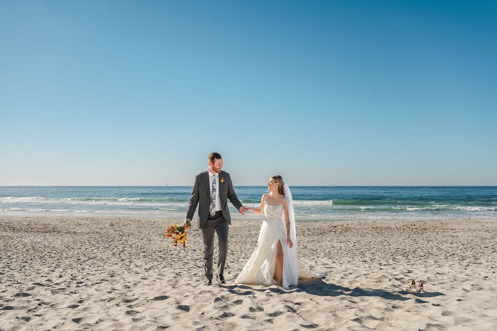 Bride and groom walk hand-in-hand on a sandy beach with ocean waves, blue sky, and a small dog nearby.