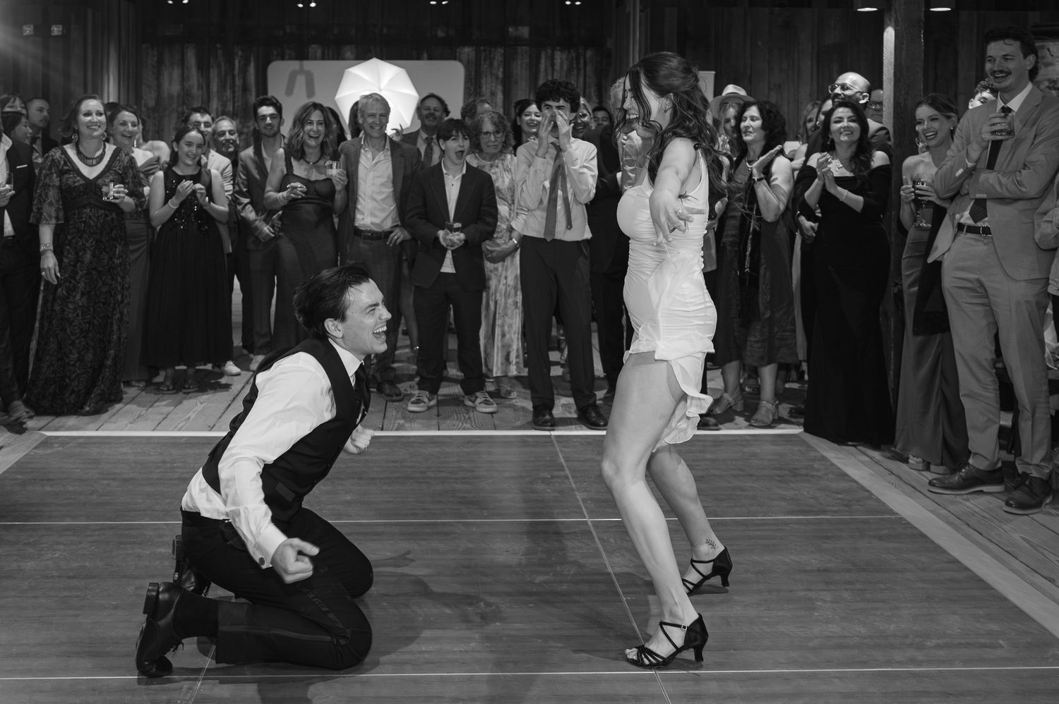 Black and white photo of a man kneeling on dance floor before a woman in a short dress, crowd watching at formal event.