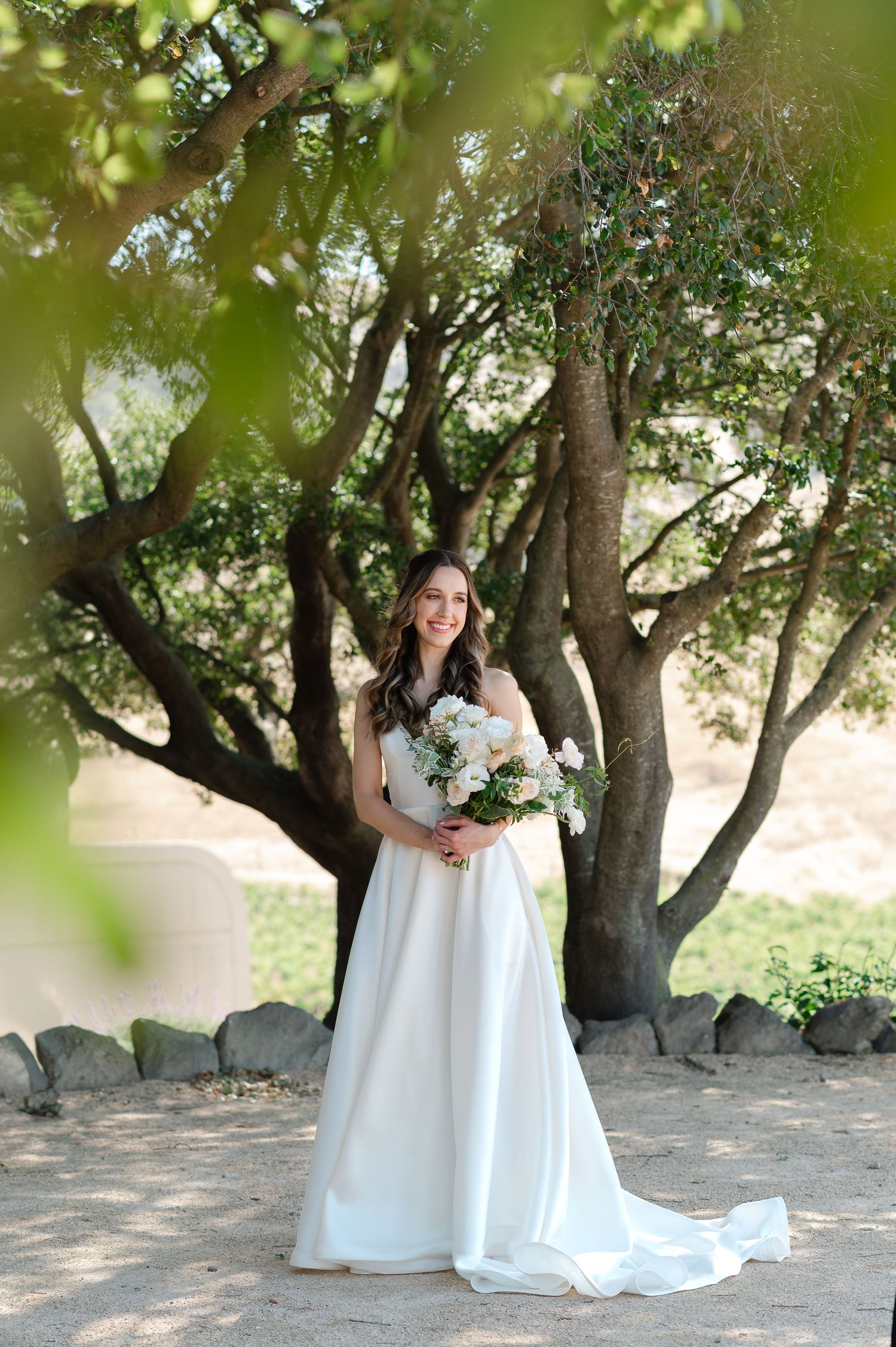 A person in a flowing white wedding dress poses under a canopy of lush green trees in a natural outdoor setting.