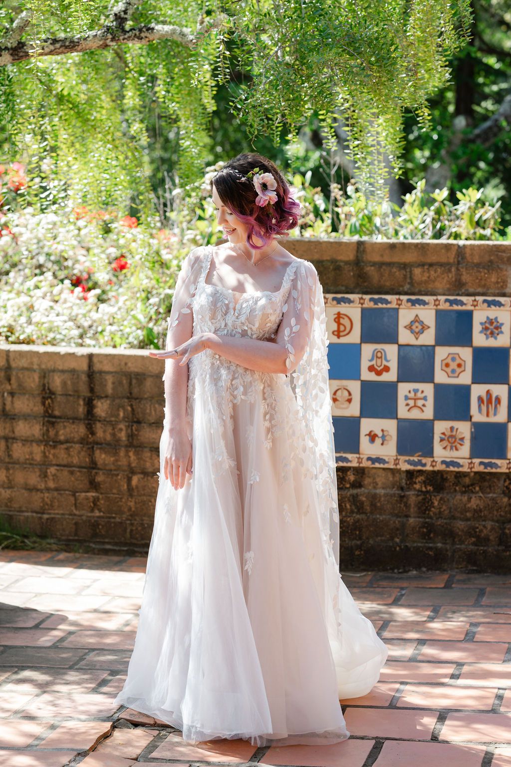 A delicate white lace wedding gown with cape photographed in a garden setting with decorative tiled wall backdrop.