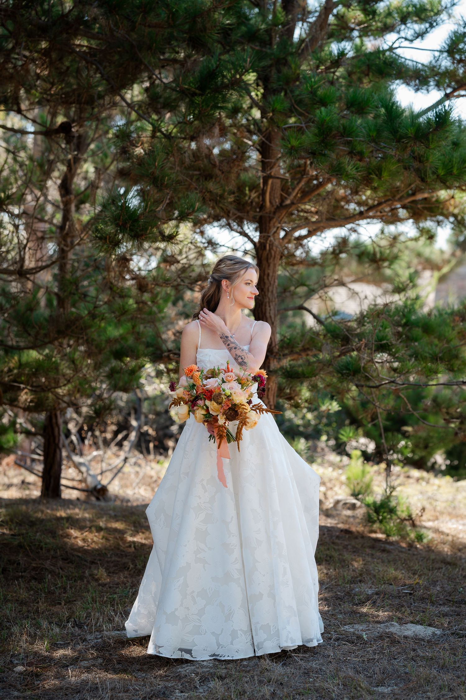 A bride in a flowing white gown holds a vibrant autumn bouquet while standing among evergreen trees in a forest setting.