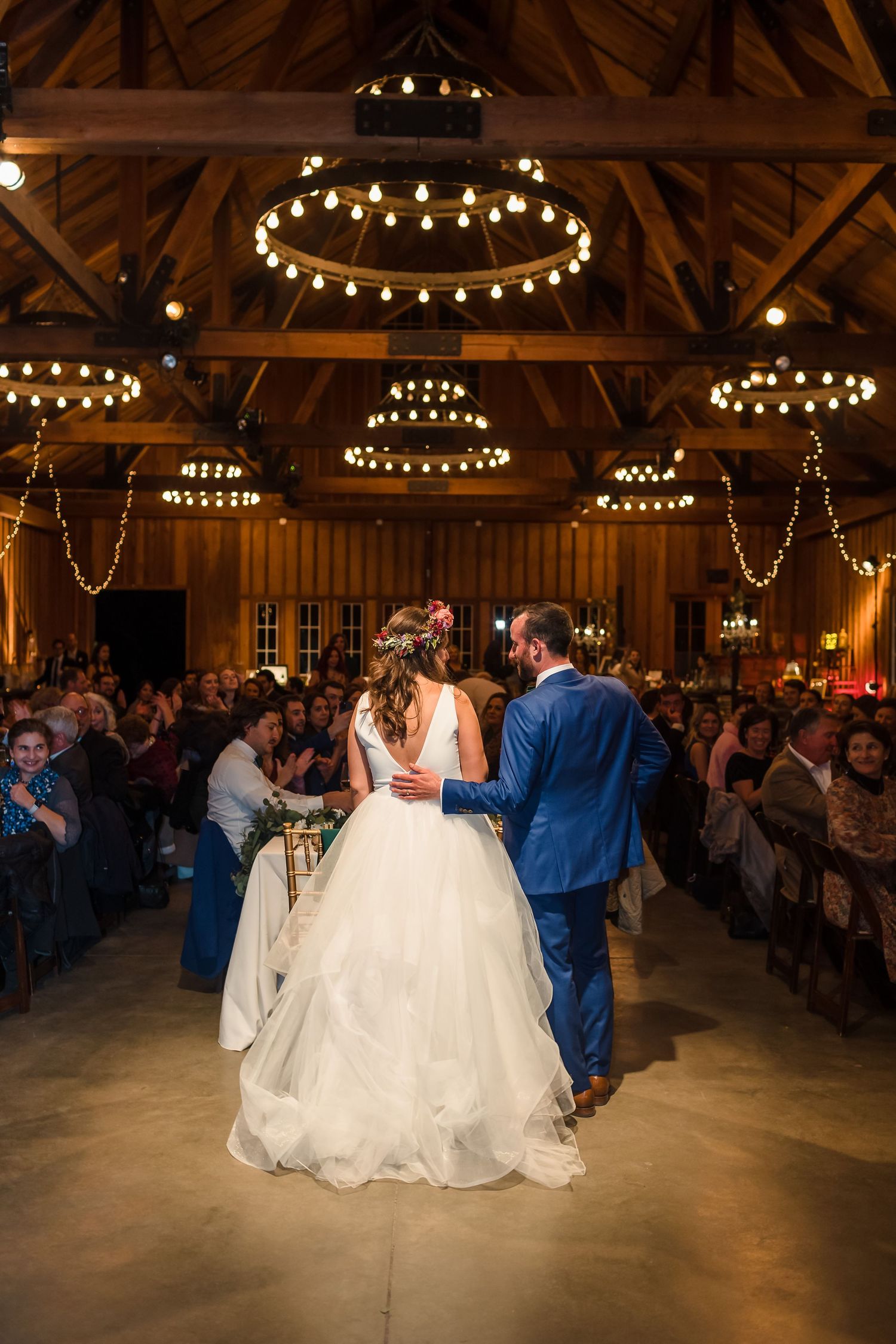 Bride and groom enter rustic barn wedding reception with string lights and chandeliers as guests watch from decorated tables.