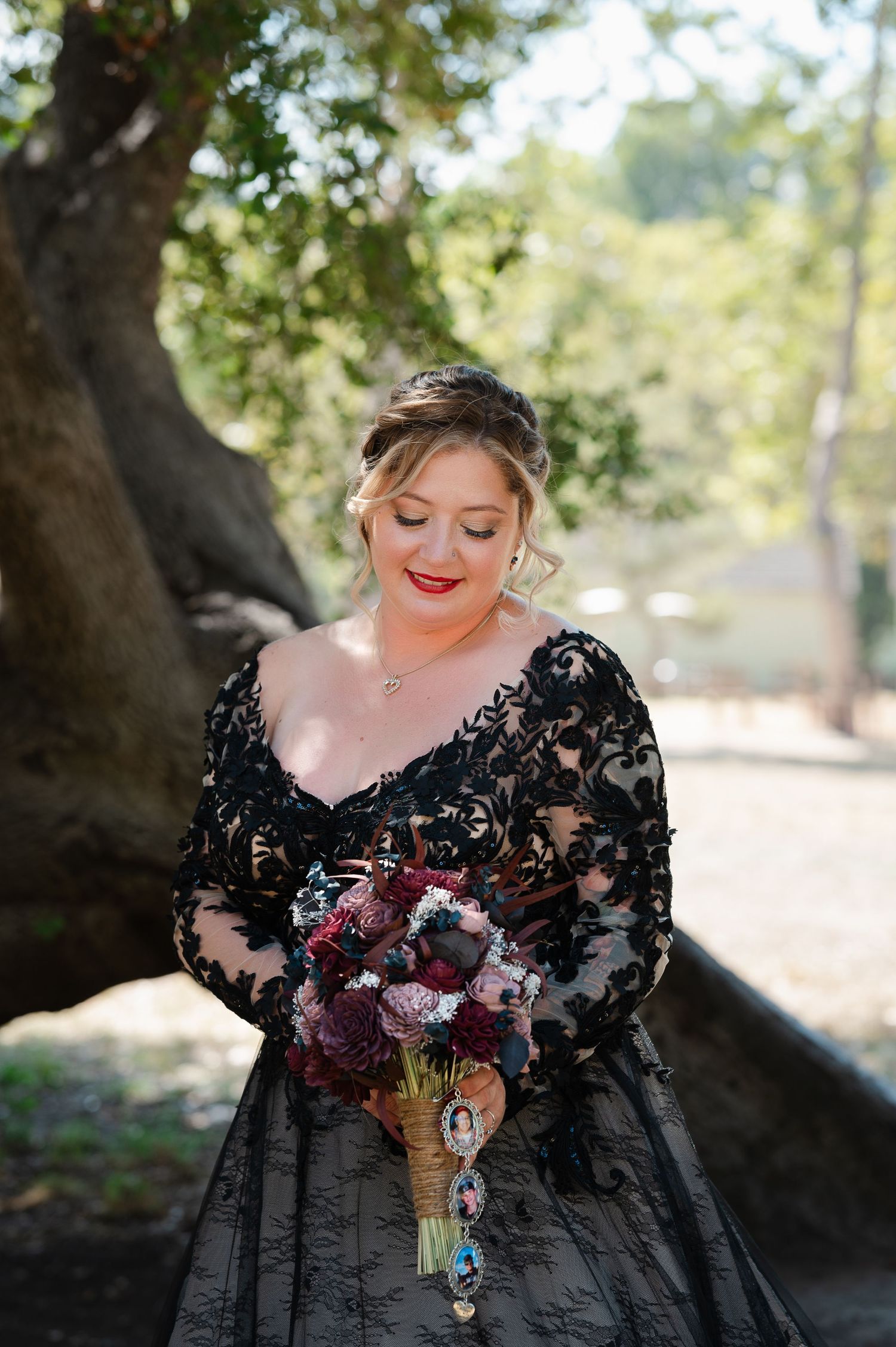 A romantic outdoor portrait in a black lace gown with deep burgundy floral bouquet against a sunlit forest backdrop.