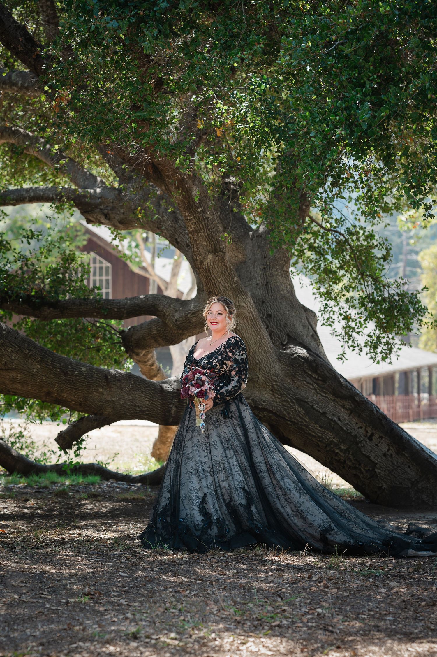 A person in an elegant black gown poses under a sprawling tree in a natural outdoor setting.