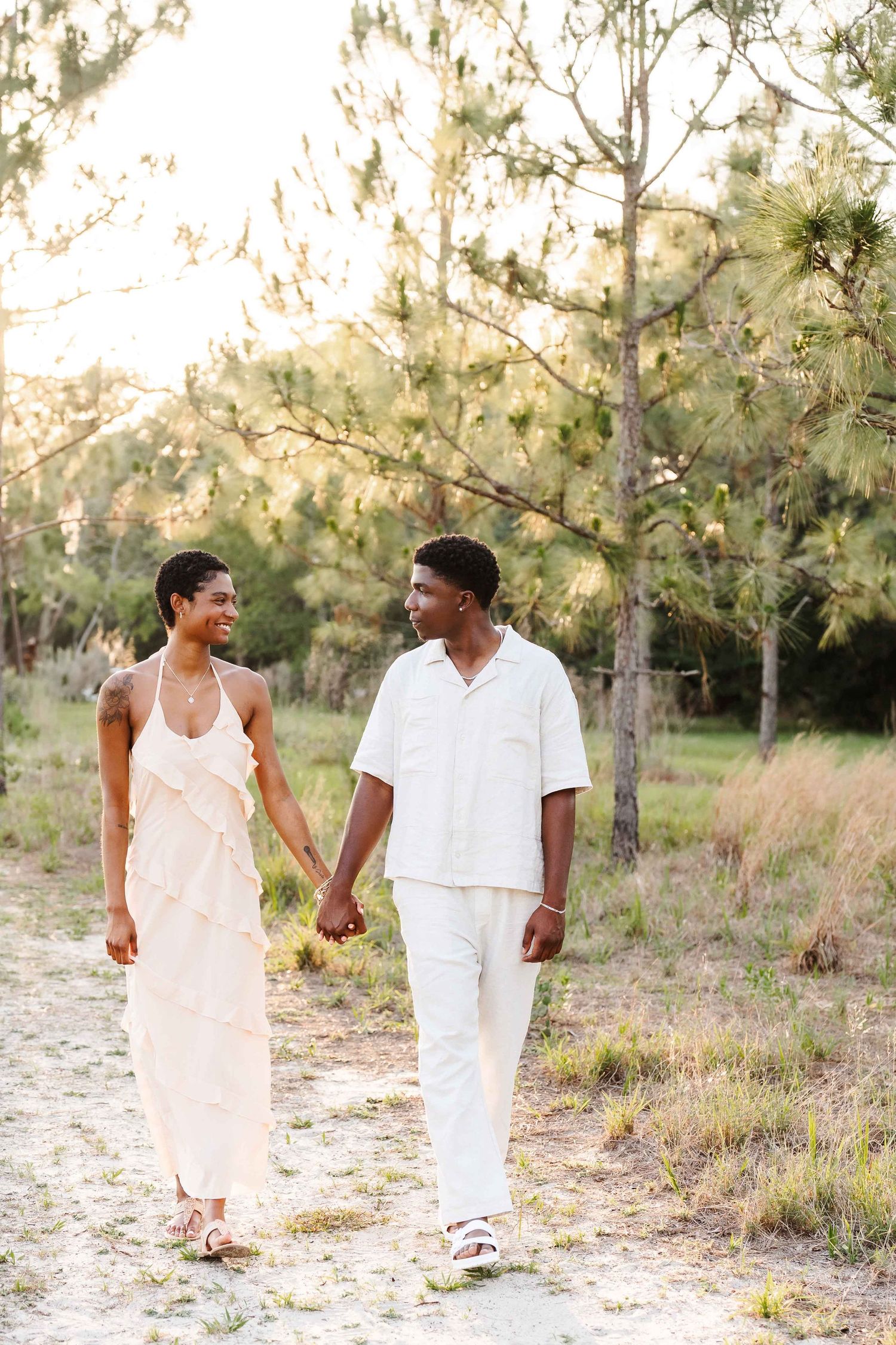 A couple in white outfits holds hands while walking on a sandy path surrounded by pine trees in golden hour light.