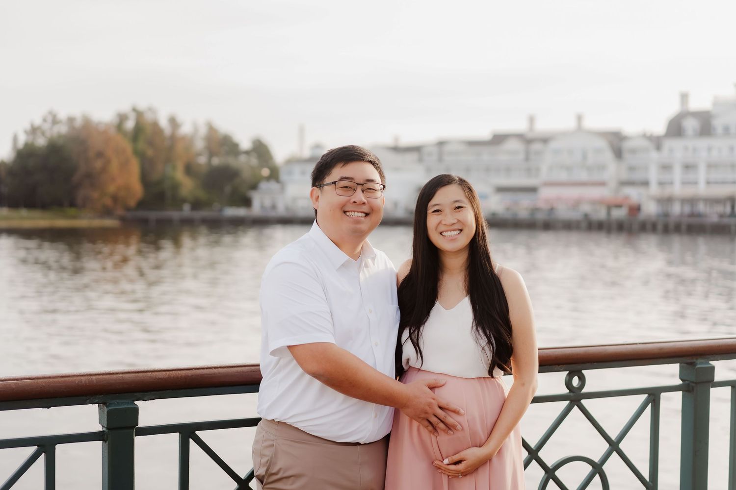 Couple posing together on a waterfront boardwalk, woman in pink skirt cradling baby bump, man in white shirt smiling.