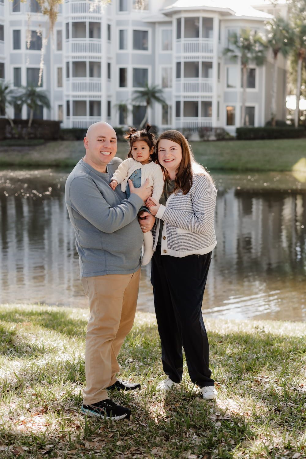 A family shares joyful moments together by a lakeside with white condominiums in the background on a sunny day.