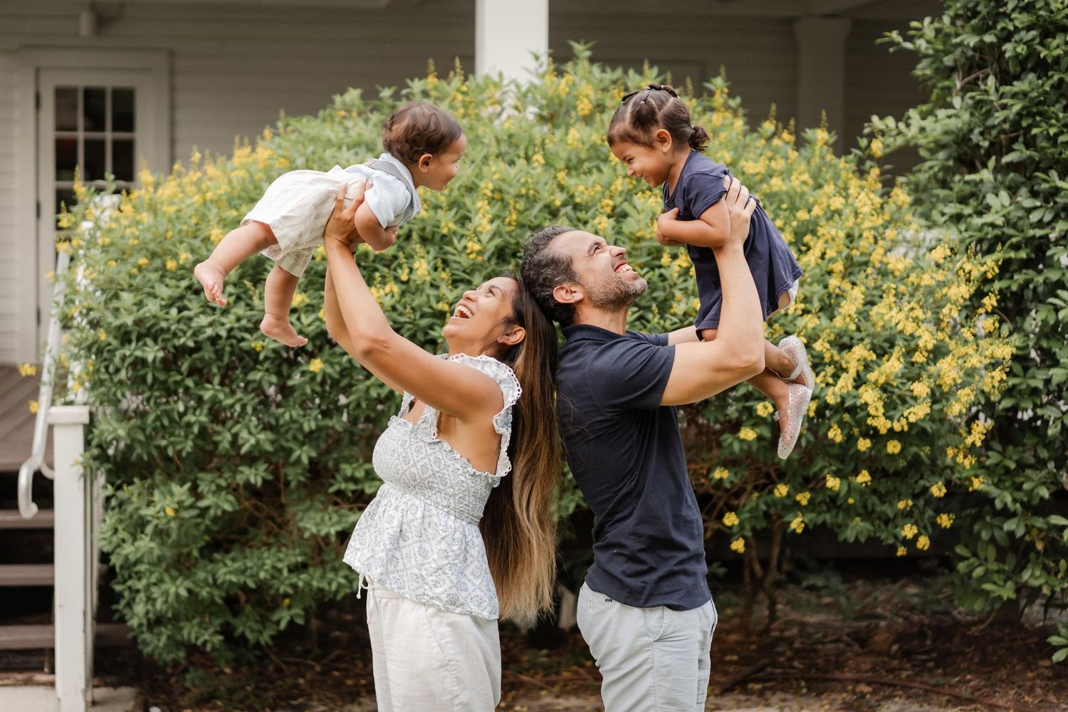 Happy family outdoors, parents lifting two young children with yellow flowering bushes in background.