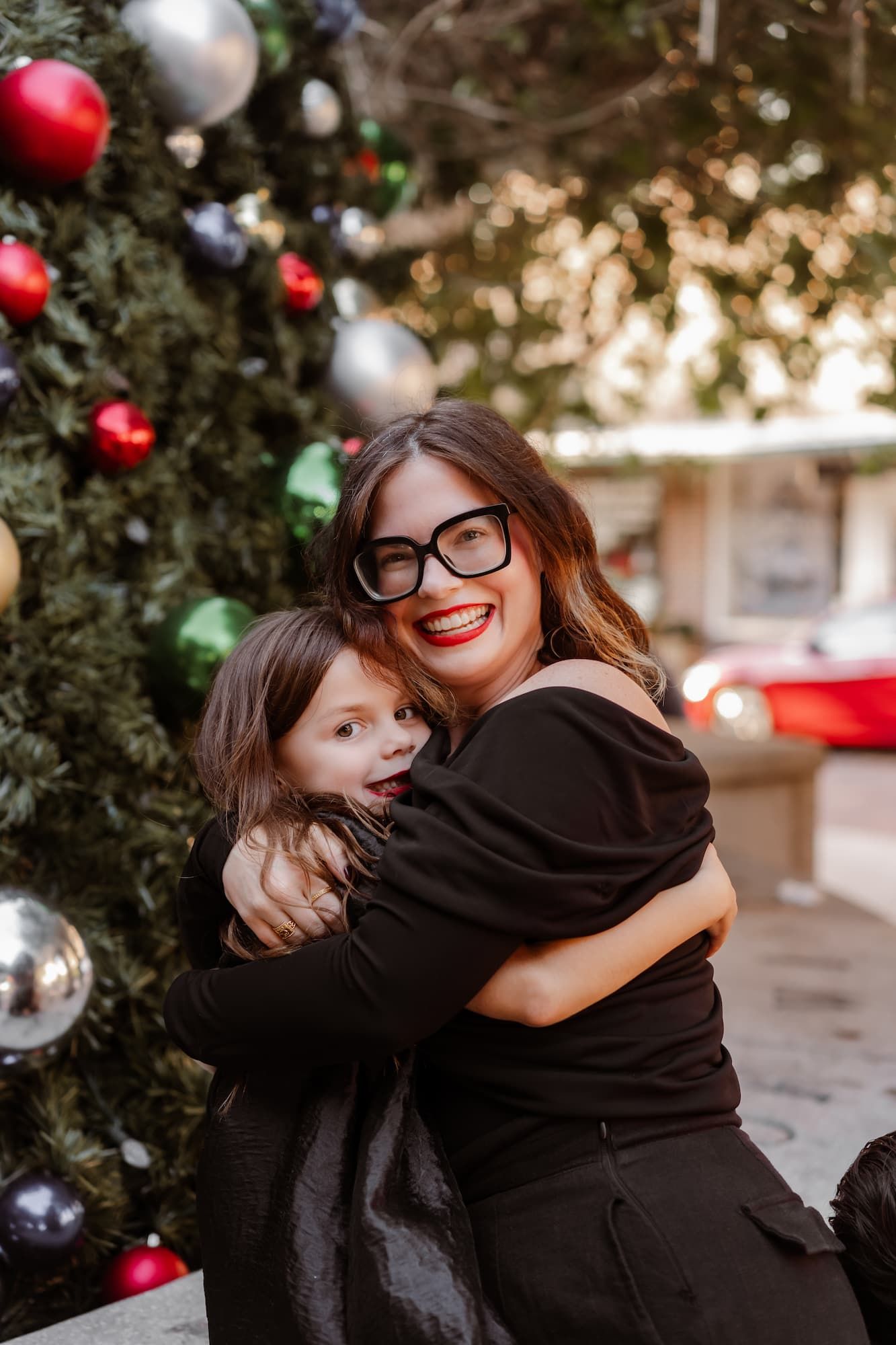 Woman in black outfit and glasses hugs young child in front of decorated Christmas tree outdoors.