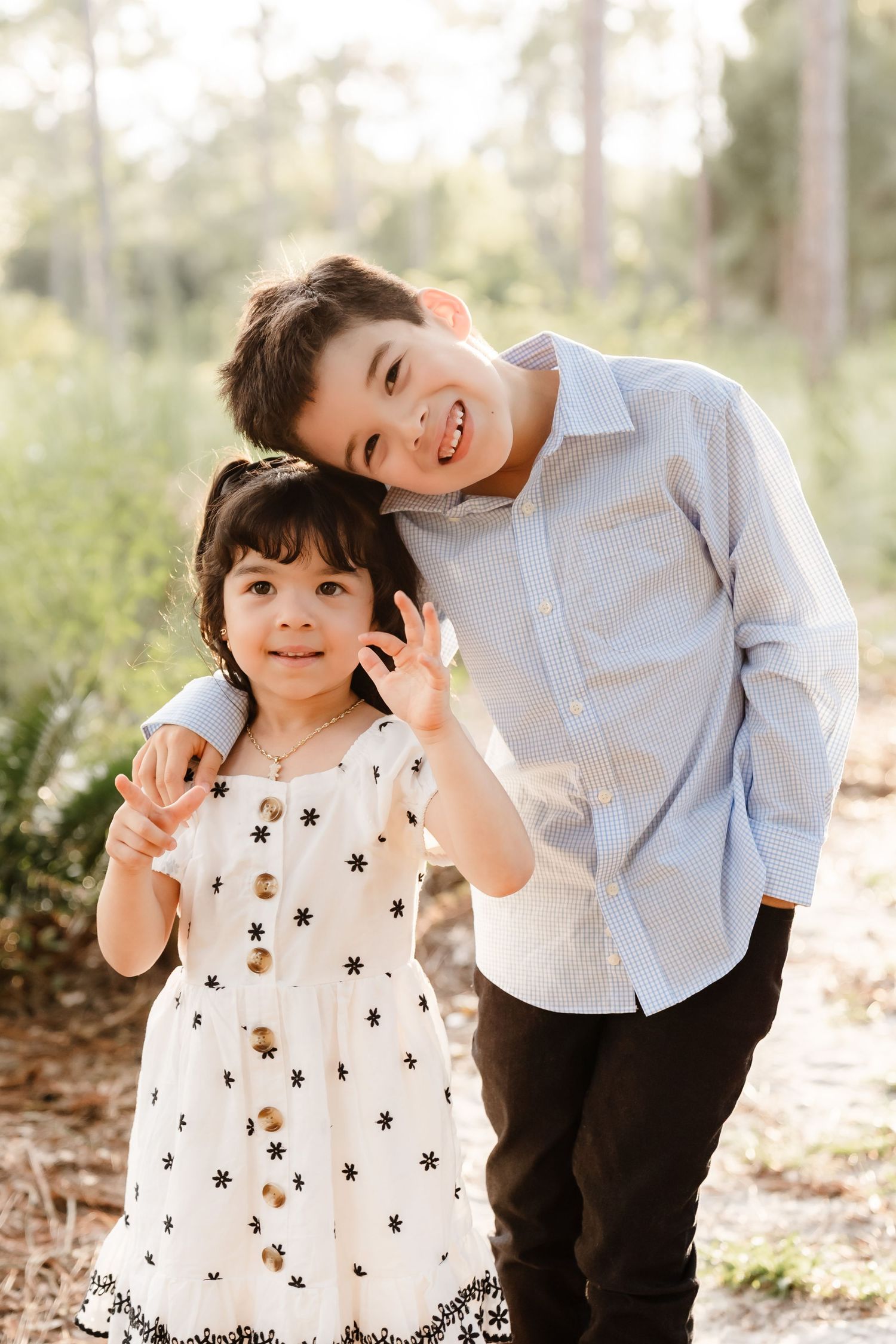 Two children in casual attire play and laugh together in a sunlit outdoor setting with trees in the background.