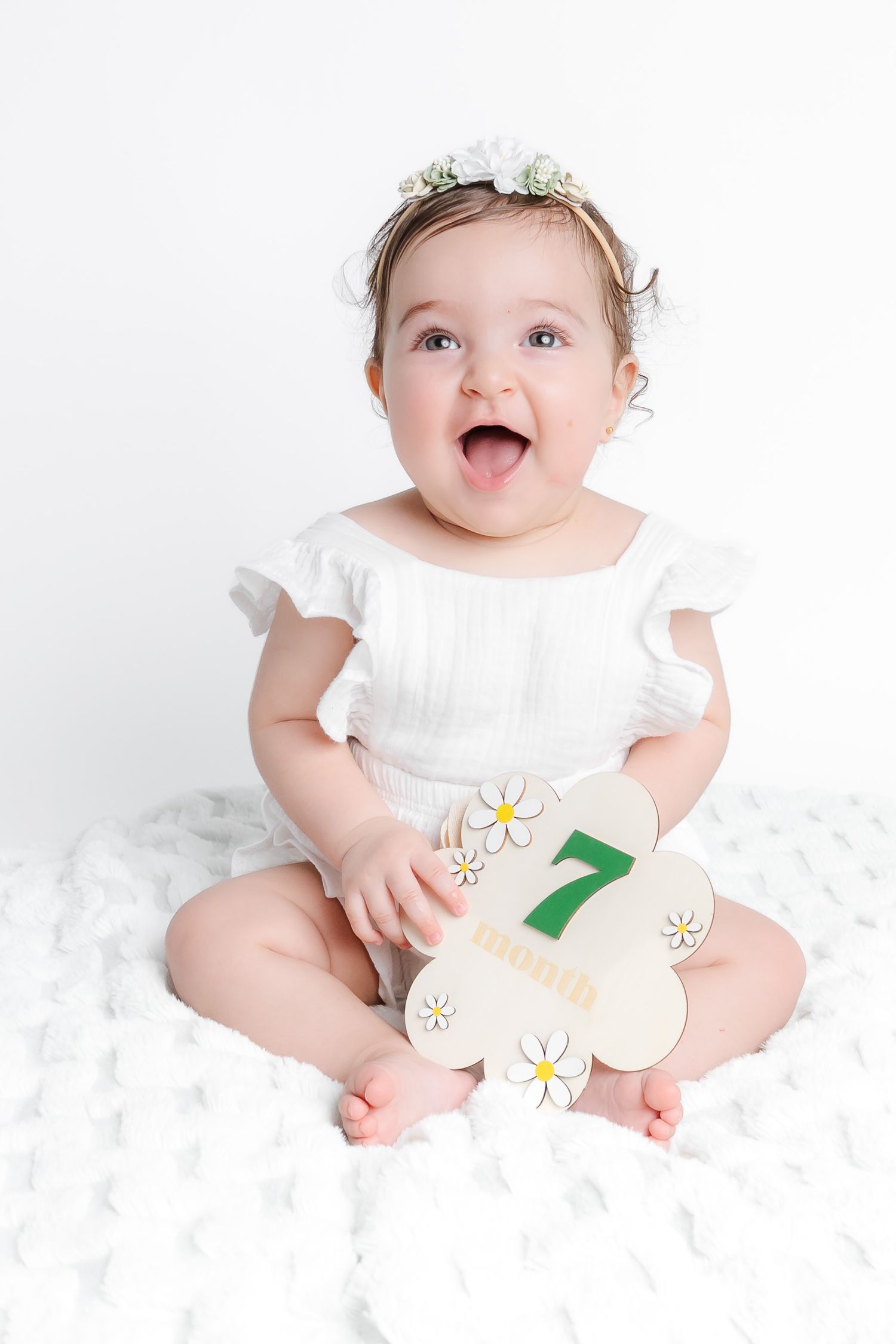 Baby wearing white outfit with daisy appliques sits on soft white background with happy expression.