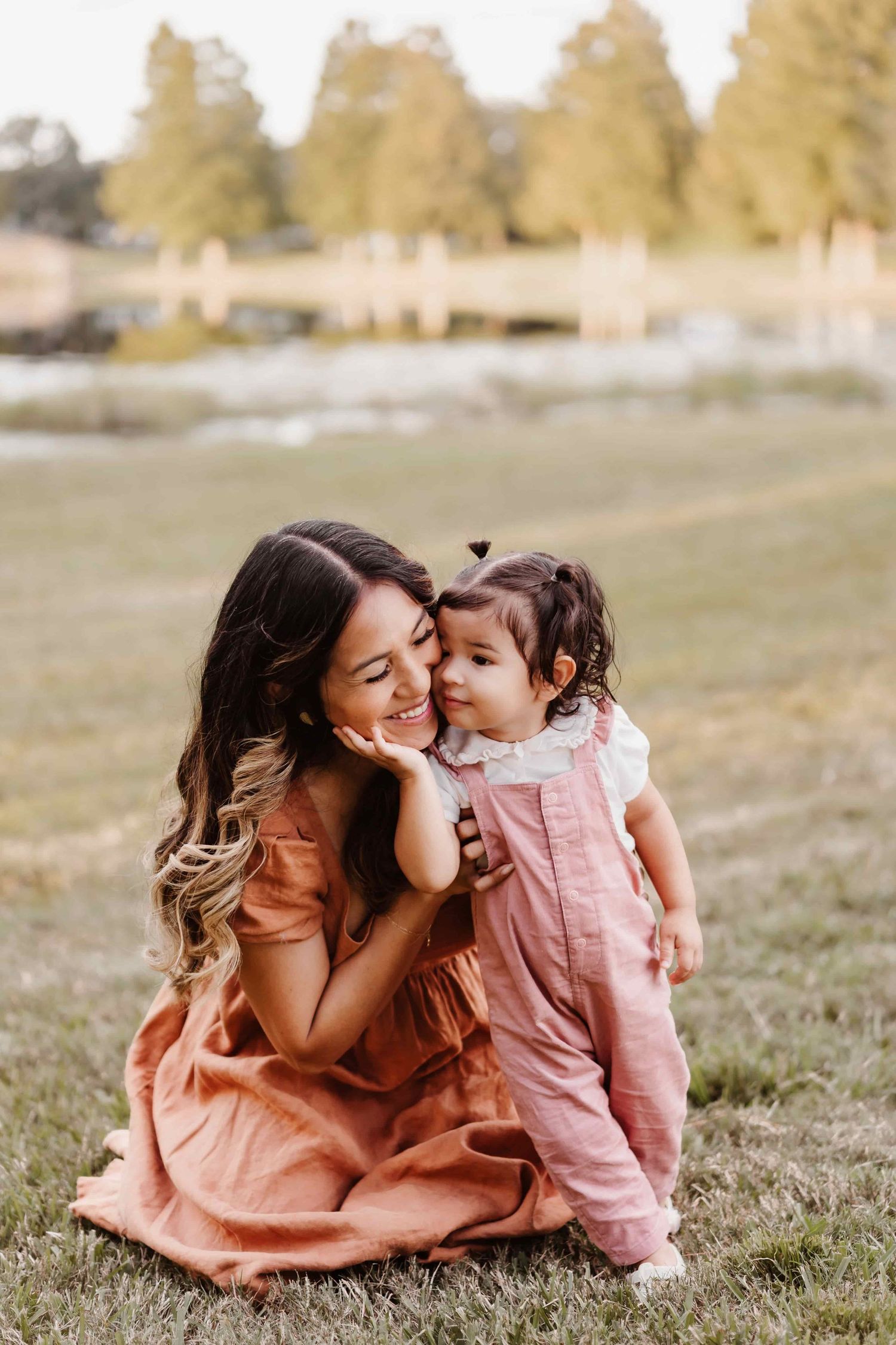A smiling mother in a pink dress sits on grass with her toddler daughter in pink overalls sharing a tender moment outdoors.