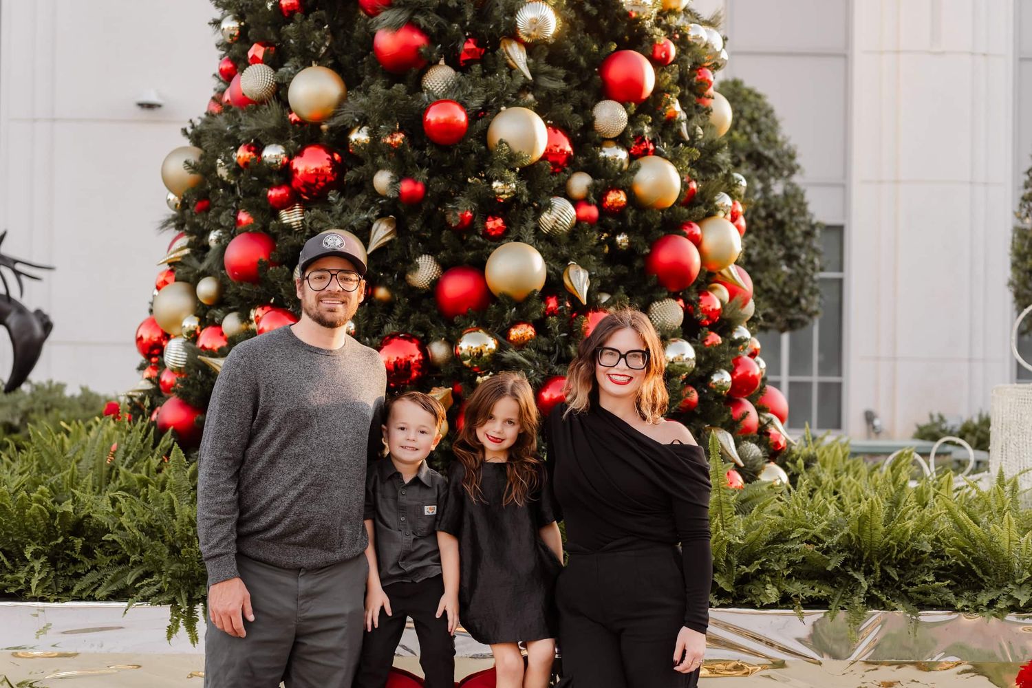 Family of four posing in front of a large decorated Christmas tree with red and gold ornaments outdoors.