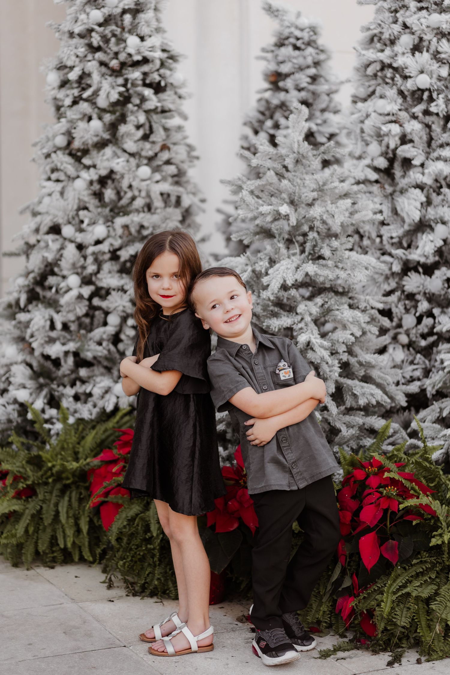 Two children in black outfits hugging in front of snow-flocked Christmas trees and red poinsettias.