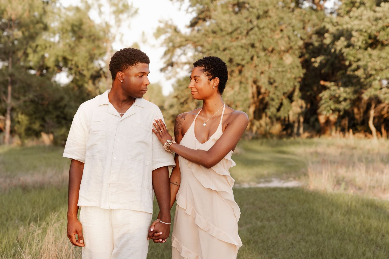 A couple in white outfits share tender moments during a romantic outdoor photoshoot in a sunlit meadow.