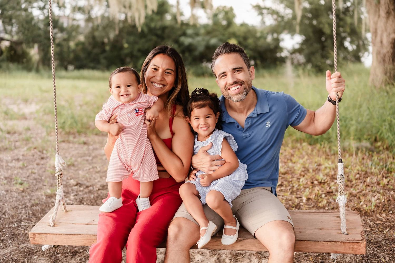 Happy family of four sitting on a wooden swing outdoors, parents holding two young daughters, smiling in a sunny park.