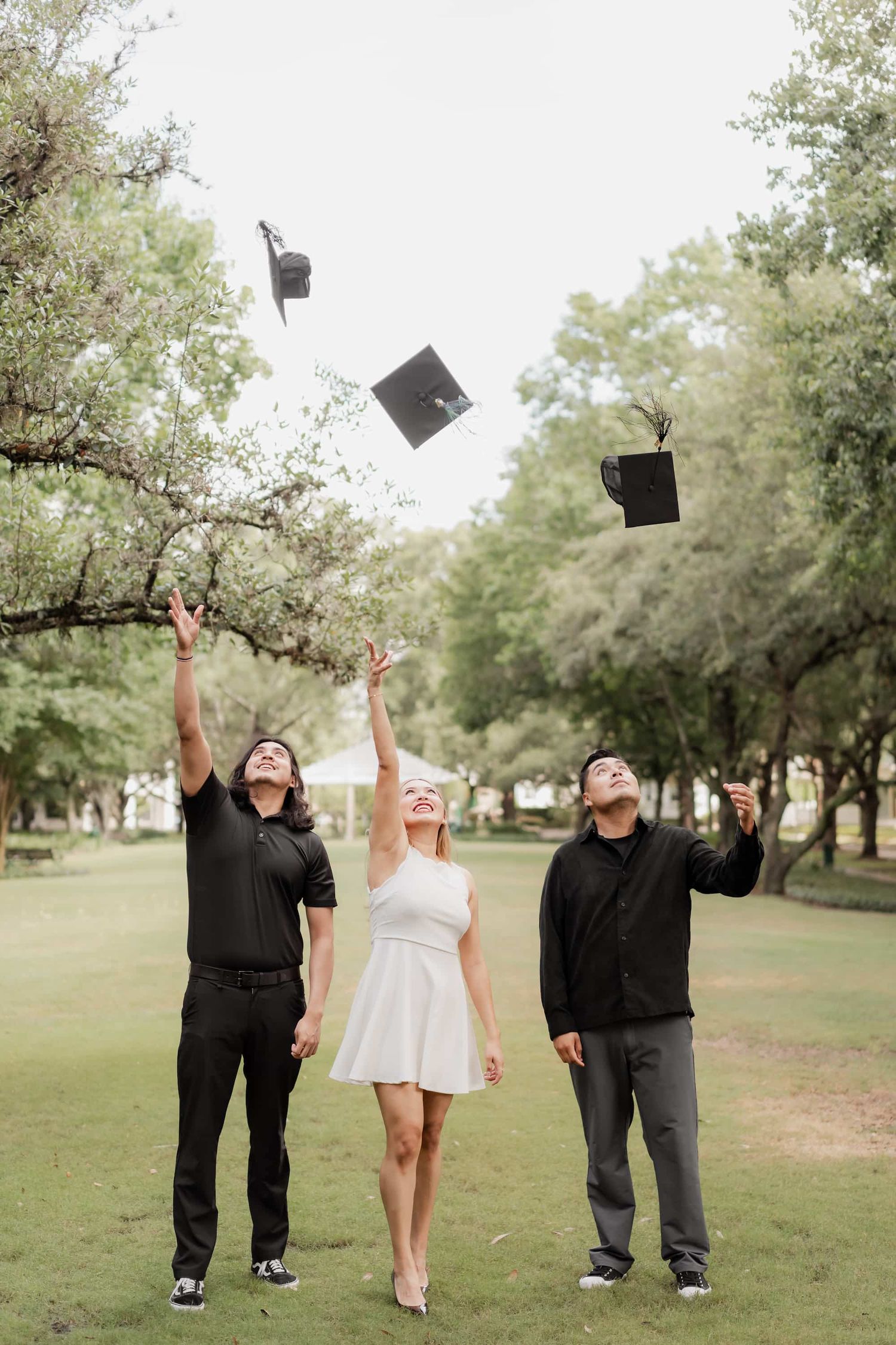 Three graduates joyfully toss their black mortarboard caps in the air outdoors in a lush green park.