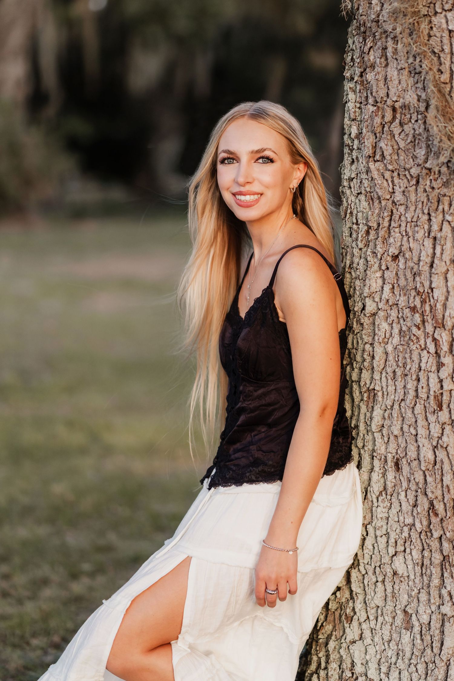 Smiling blonde woman in black lace top and white skirt leaning against a tree in a sunlit park.