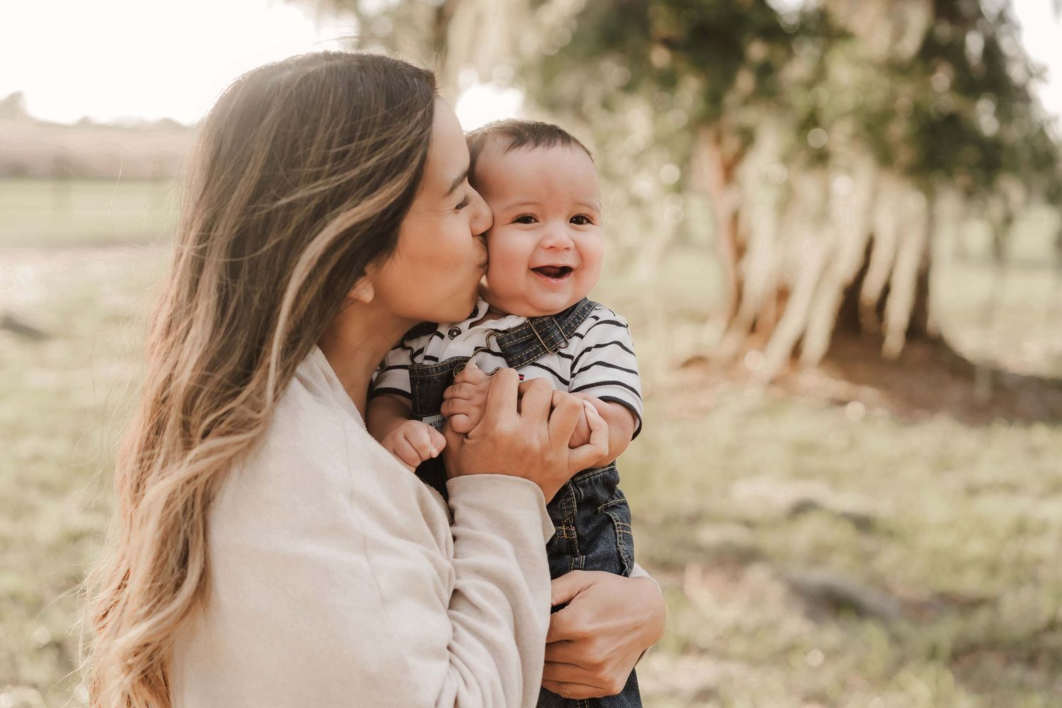 A mother kissing her smiling baby boy outdoors in a lush green park setting.