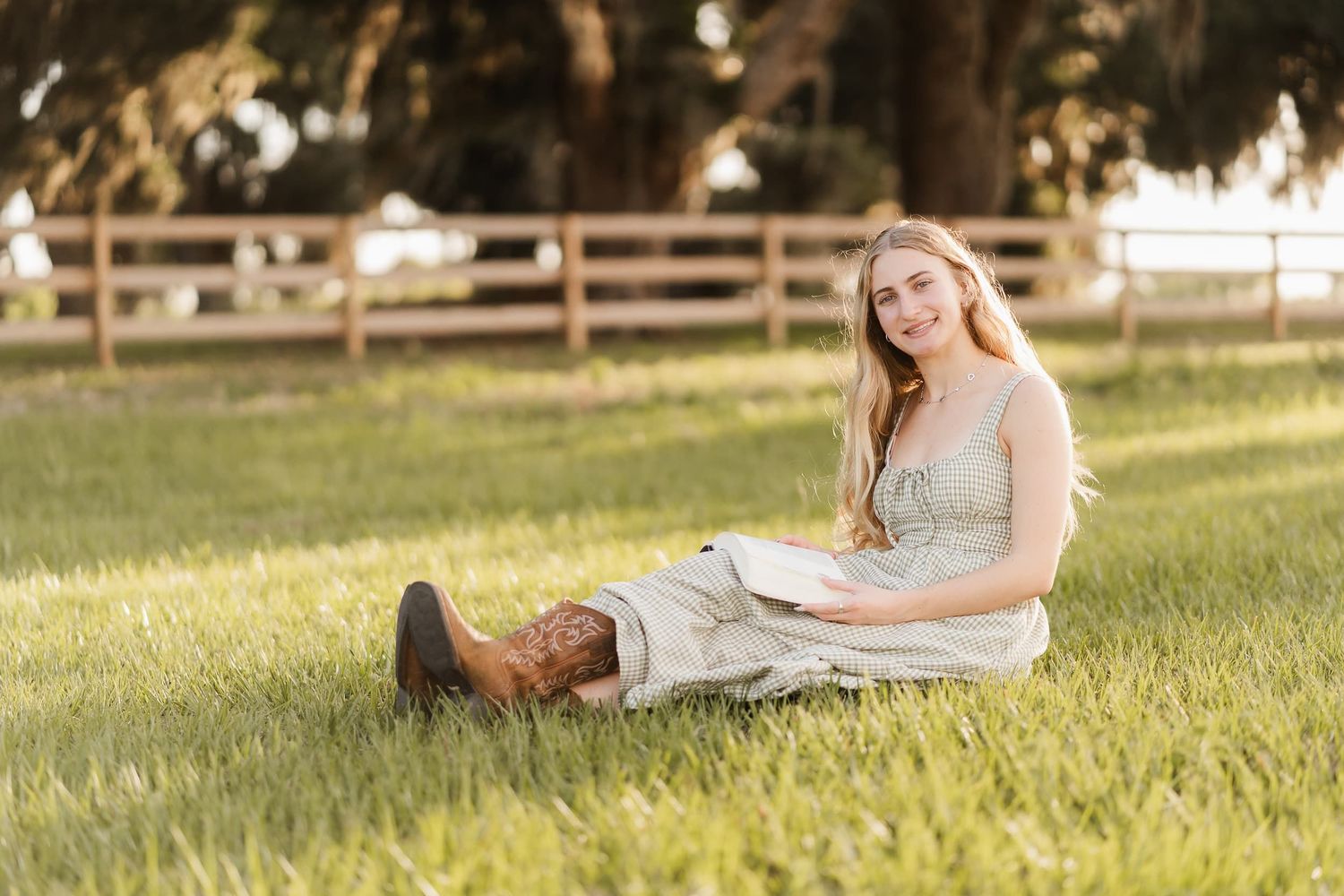 Young woman in a light dress and cowboy boots sitting on green grass with a wooden fence backdrop.