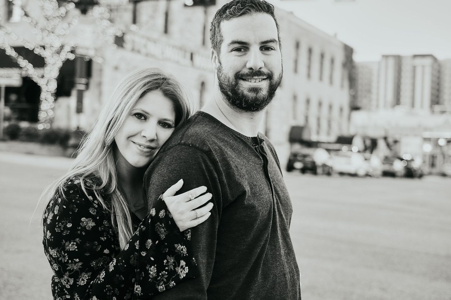 A smiling couple poses outdoors in a black and white engagement photo on a city street.