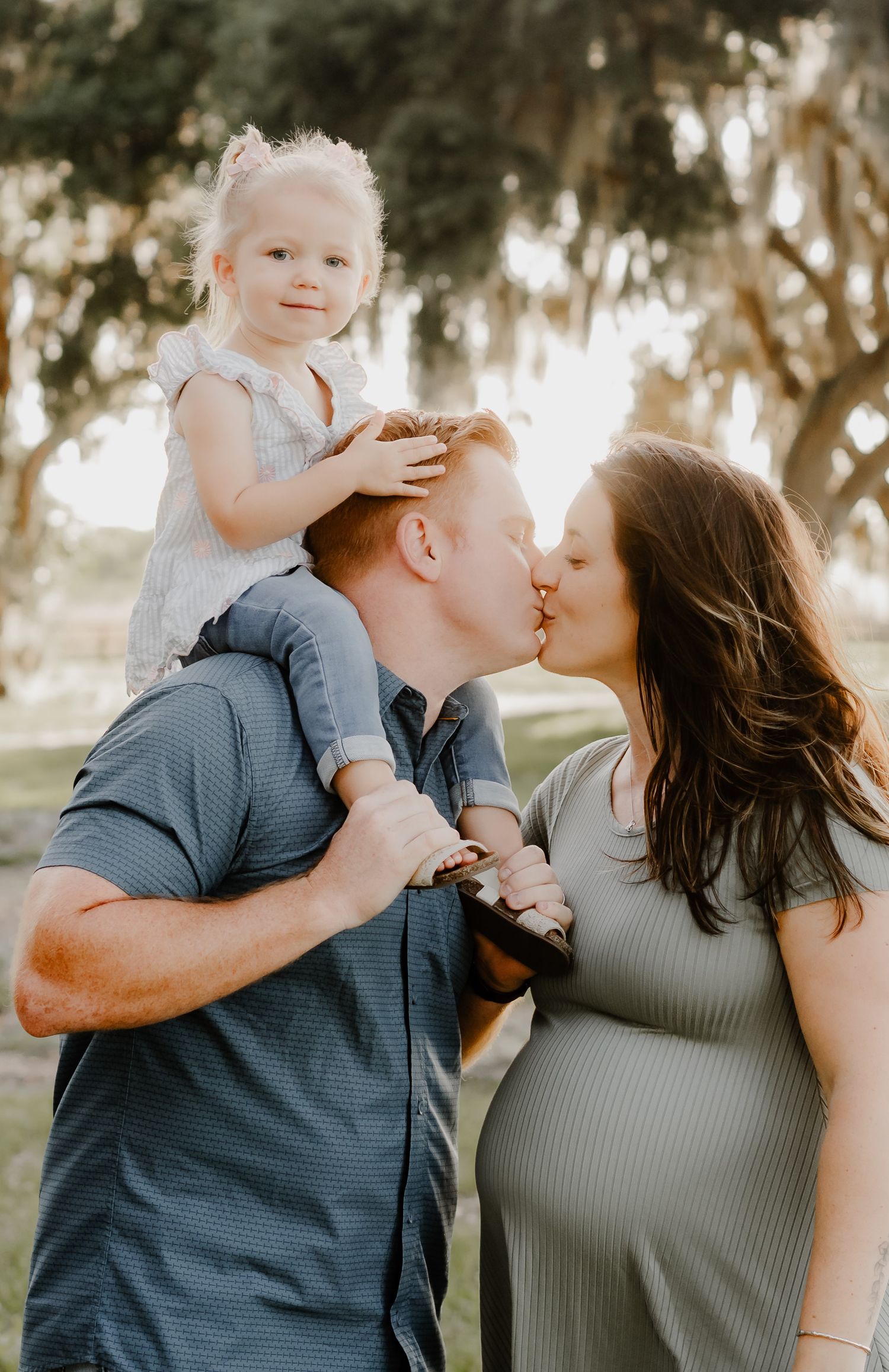 A couple kisses while their toddler daughter sits on dad's shoulders during an outdoor maternity photo session.