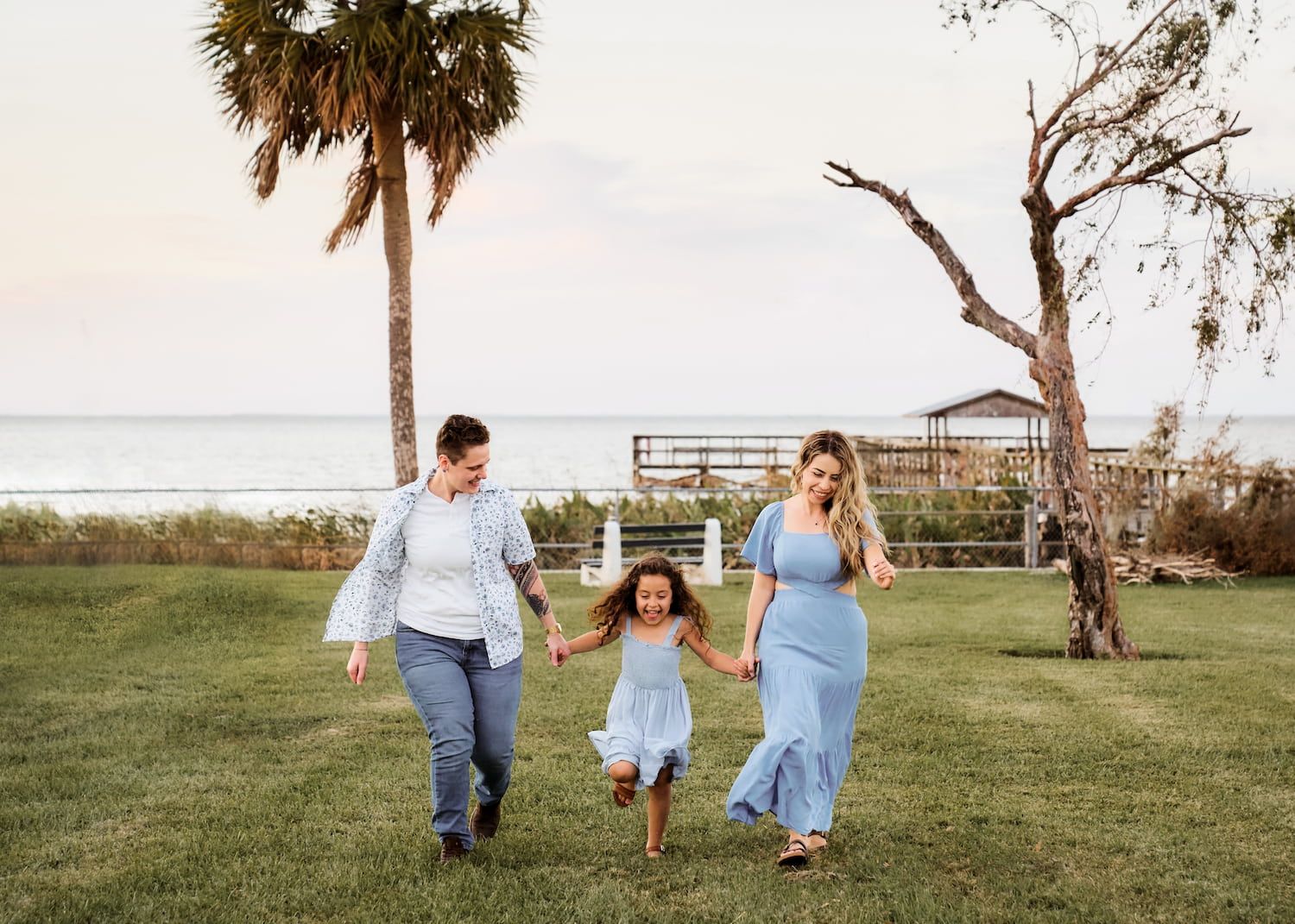 Two women and a young girl hold hands walking on grass near a beach with palm trees in soft evening light.