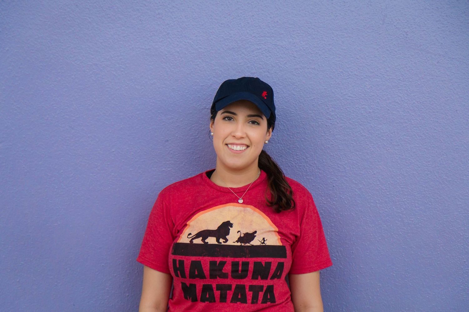 Smiling woman wearing a red Hakuna Matata Lion King t-shirt and blue cap against a purple wall.