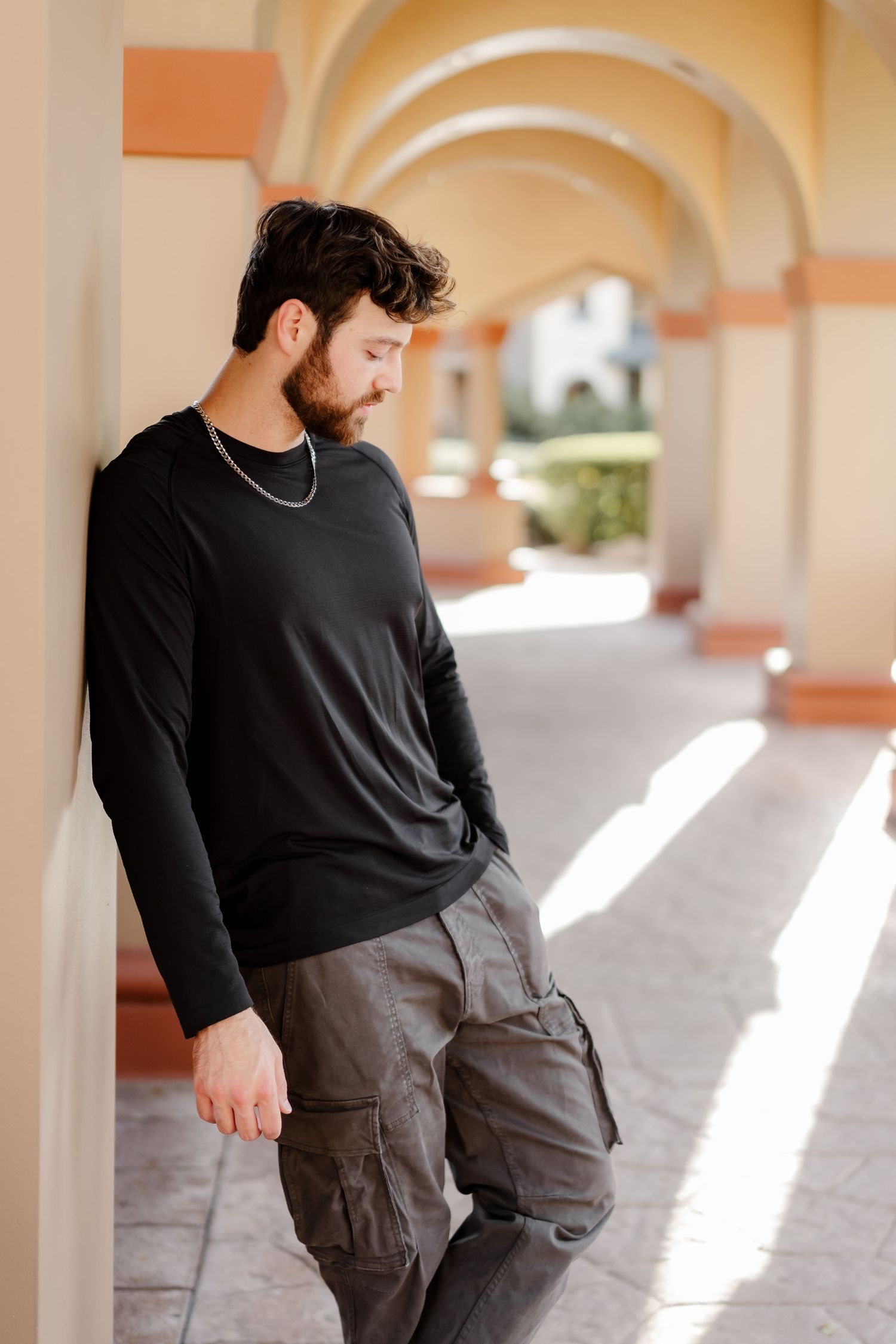 Bearded man in black long-sleeve shirt and cargo pants leaning against a pillar in a sunlit arched corridor.
