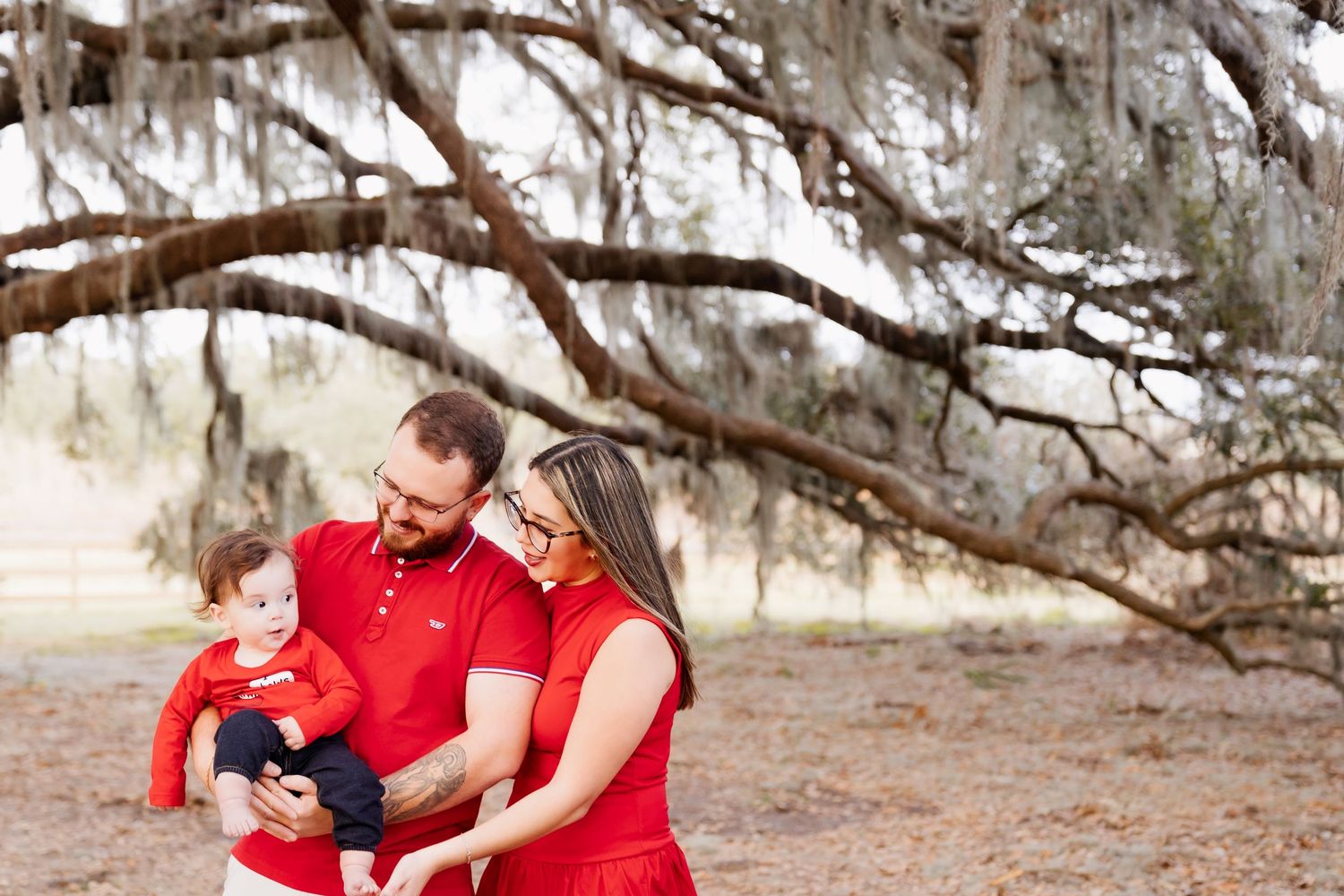 Family of three in matching red outfits under a large oak tree with Spanish moss, parents lovingly holding their baby.