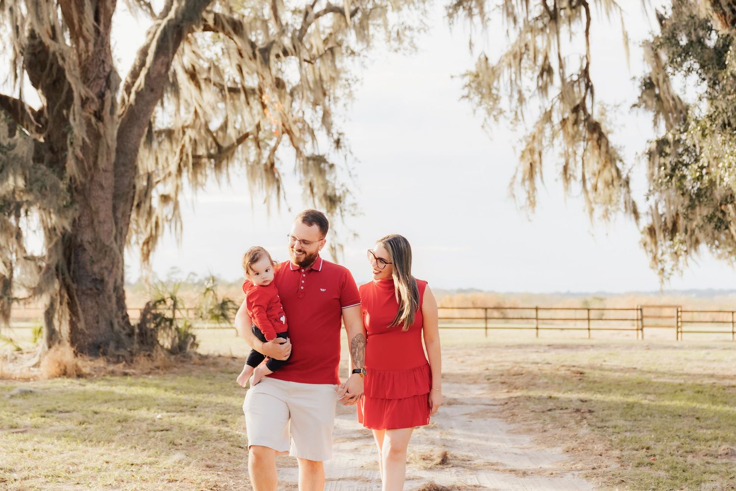 Family of three walking on a sunny dirt path under a moss-draped oak tree, wearing coordinating red outfits.