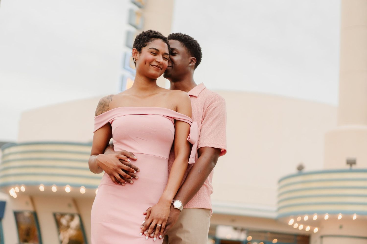 A couple in pink outfits shares a romantic moment outdoors, with the man kissing the woman's cheek.