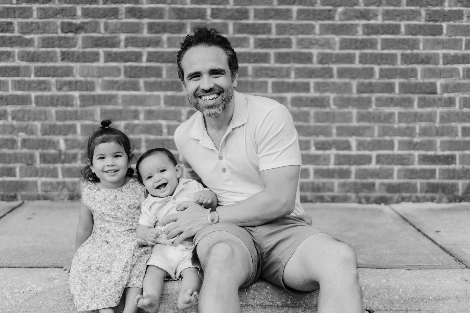 Black and white photo of a smiling man sitting on steps with two young children in front of a brick wall.