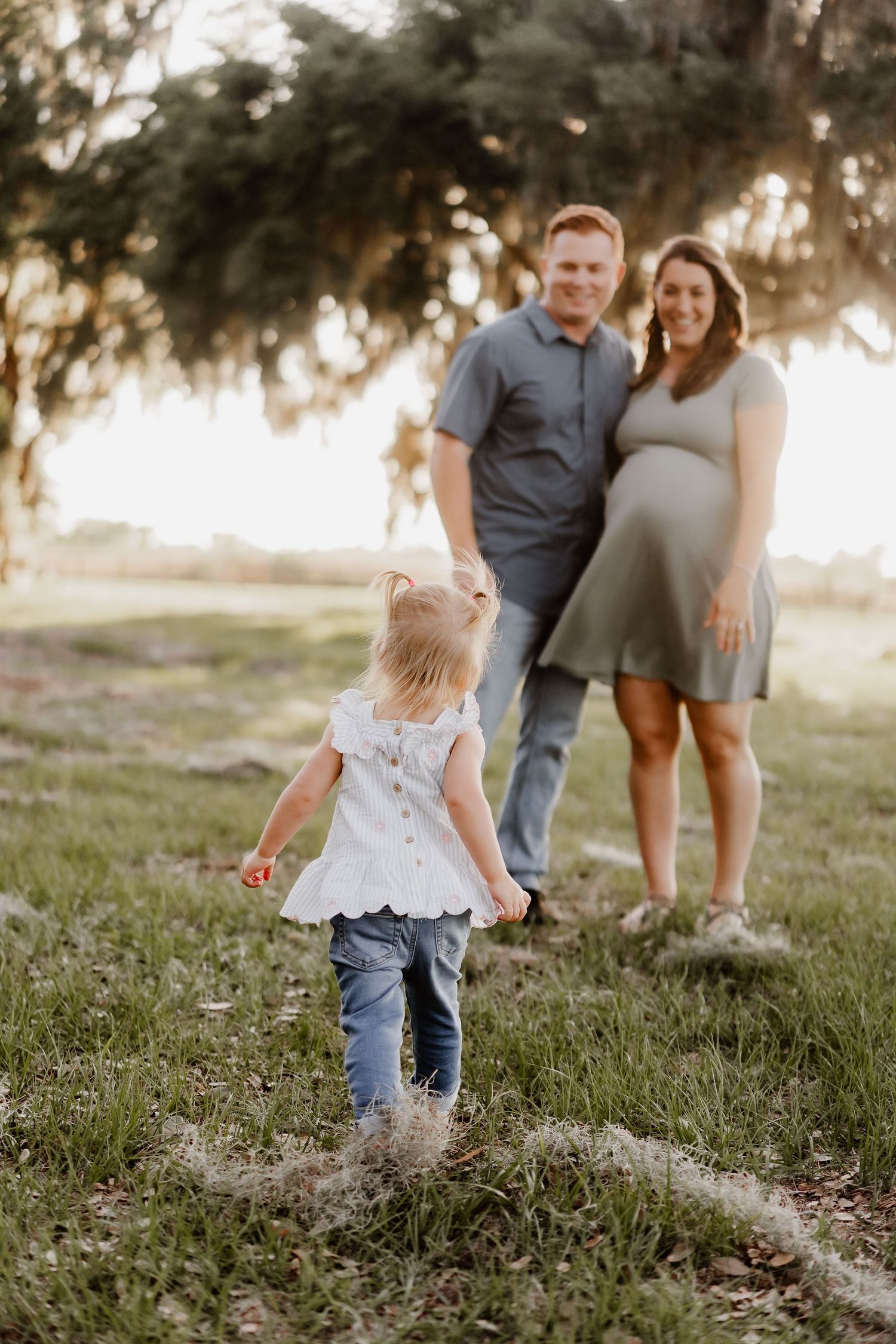 A toddler girl walks toward her pregnant mother and father during a golden hour family maternity photo session outdoors.