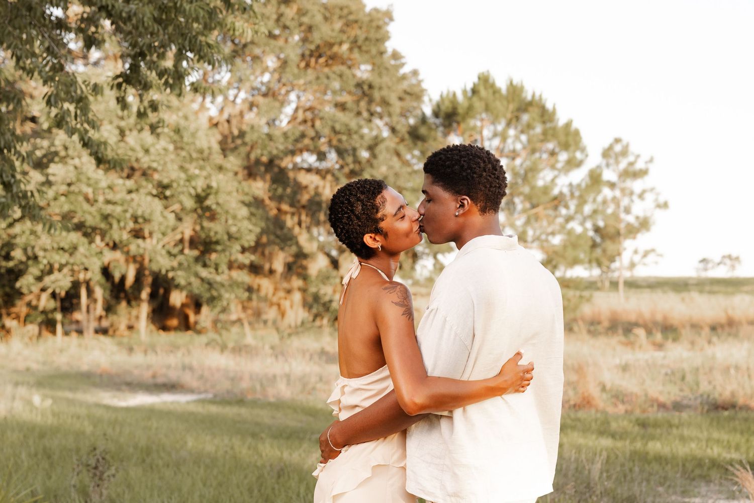 A couple shares a romantic kiss outdoors in a lush green field, dressed in elegant white and blush attire.