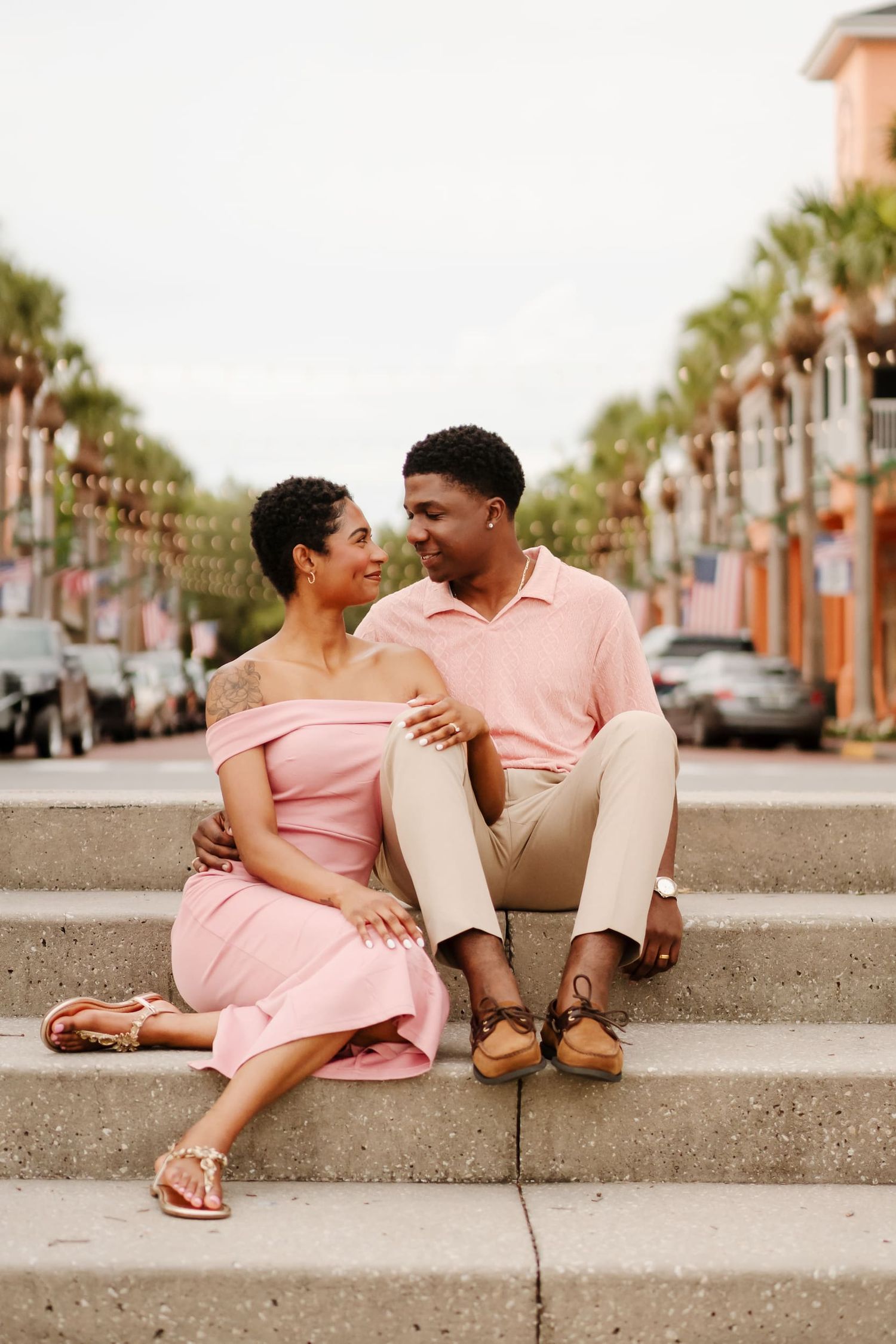 A couple in coordinating pink outfits sits on outdoor steps, gazing at each other with palm trees in the background.