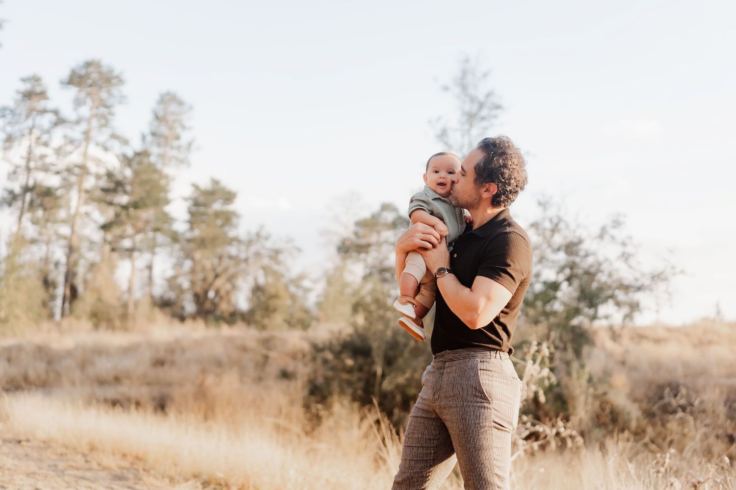 Parent and baby share a tender moment outdoors in warm sunlight with pine trees in background.