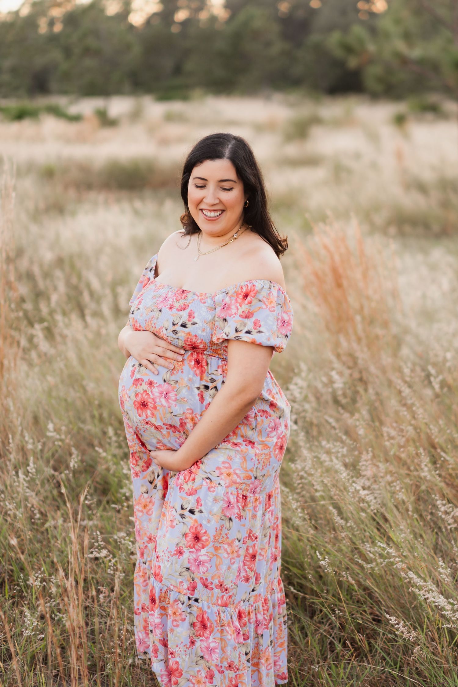 A person in a pink floral off-shoulder dress poses gracefully in a sunlit field of tall grass during golden hour.