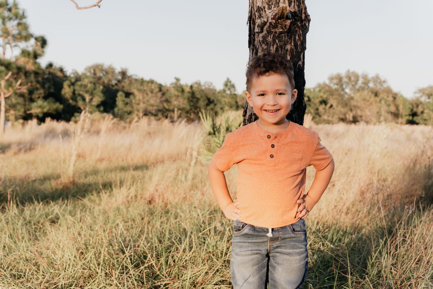 A child in an orange shirt and jeans poses in a grassy field during golden hour.