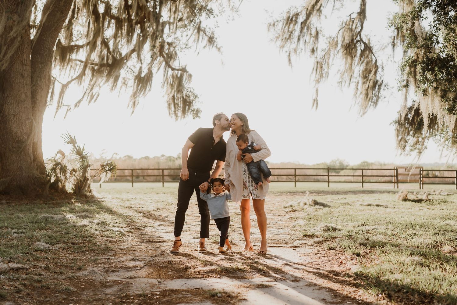 Family sharing a tender moment on a sunlit farm path under Spanish moss-draped oak trees.