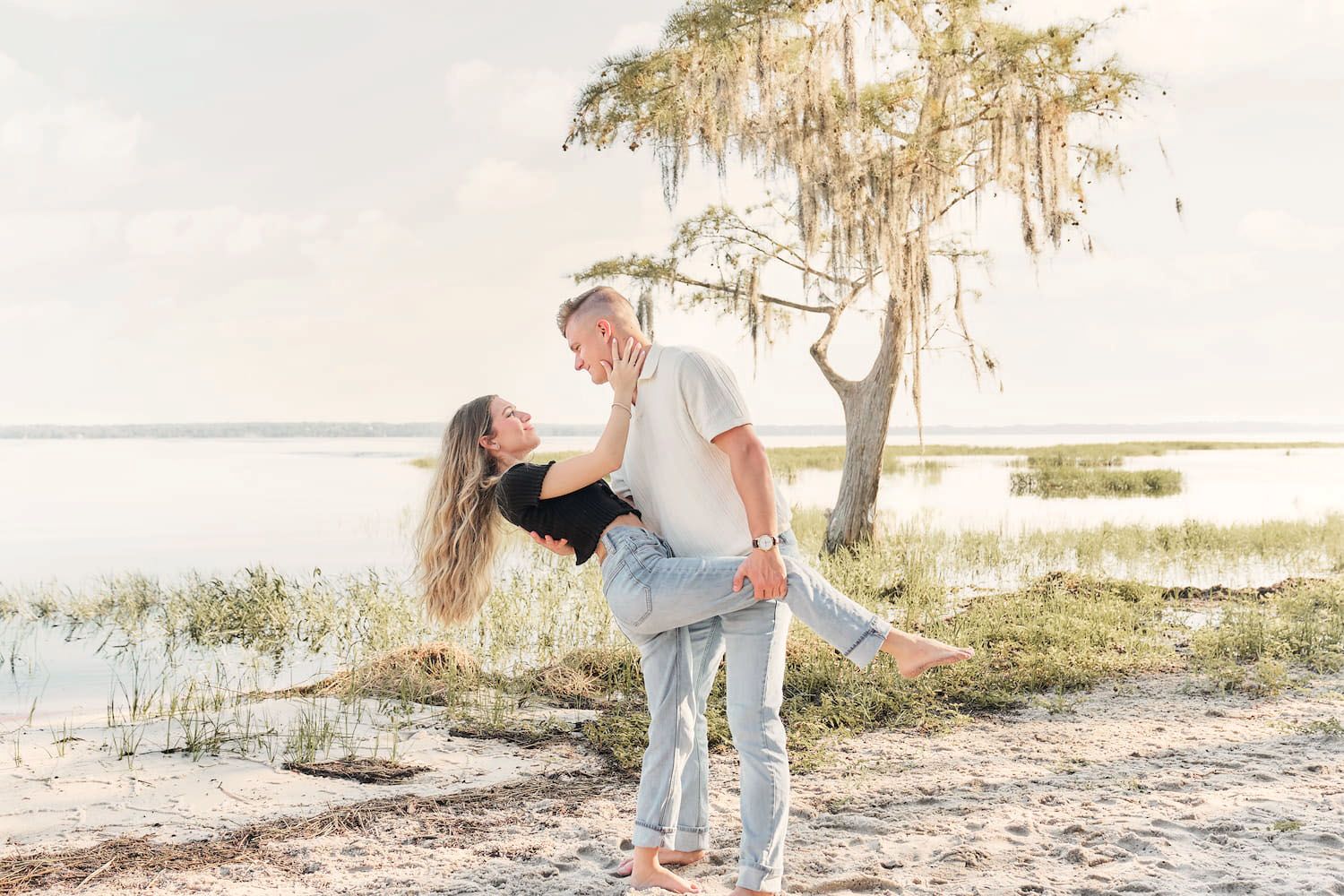 Couple sharing a romantic lift embrace on a sandy lakeside beach with Spanish moss trees at golden hour.