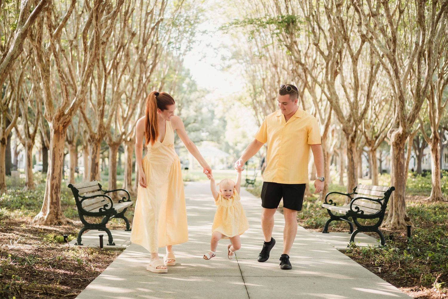 A happy family in yellow outfits walks along a sunny tree-lined path, parents holding their toddler's hands.