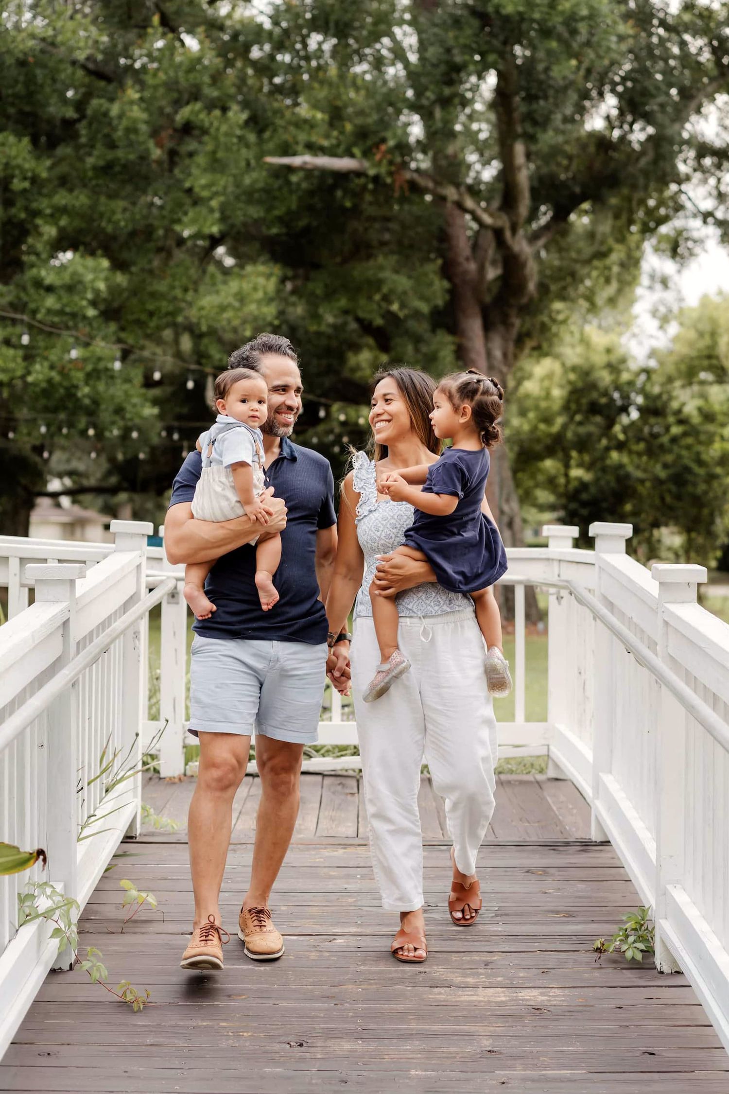 A happy family of four walks on a white wooden bridge surrounded by lush green trees.