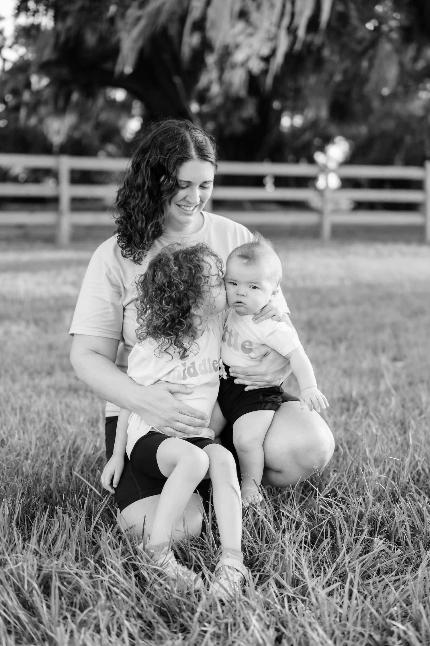 Black and white photo of a smiling mother kneeling in grass with two young children on a sunny day near a wooden fence.