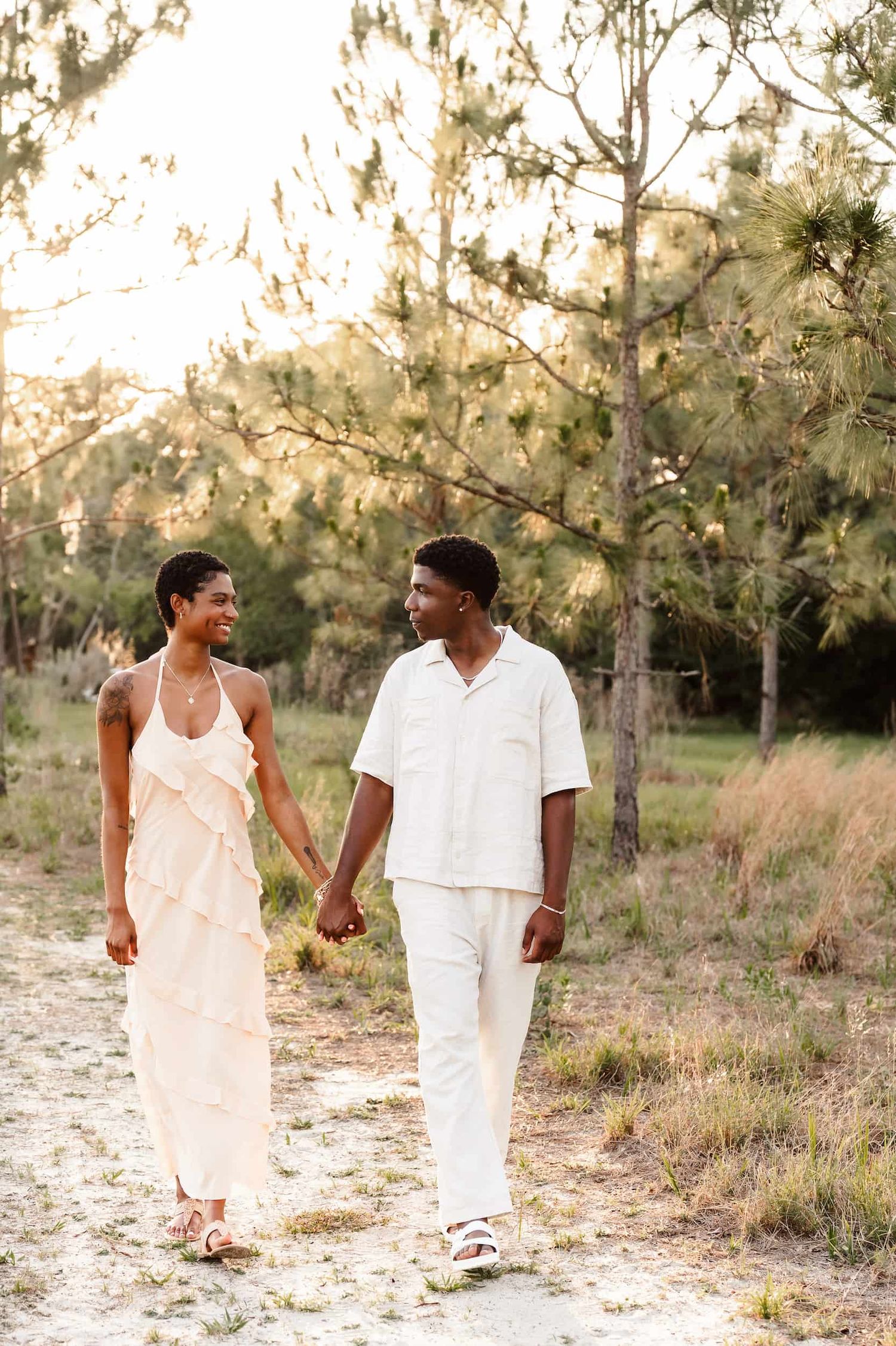 A couple in white outfits walking and holding hands along a sandy path surrounded by trees at sunset.