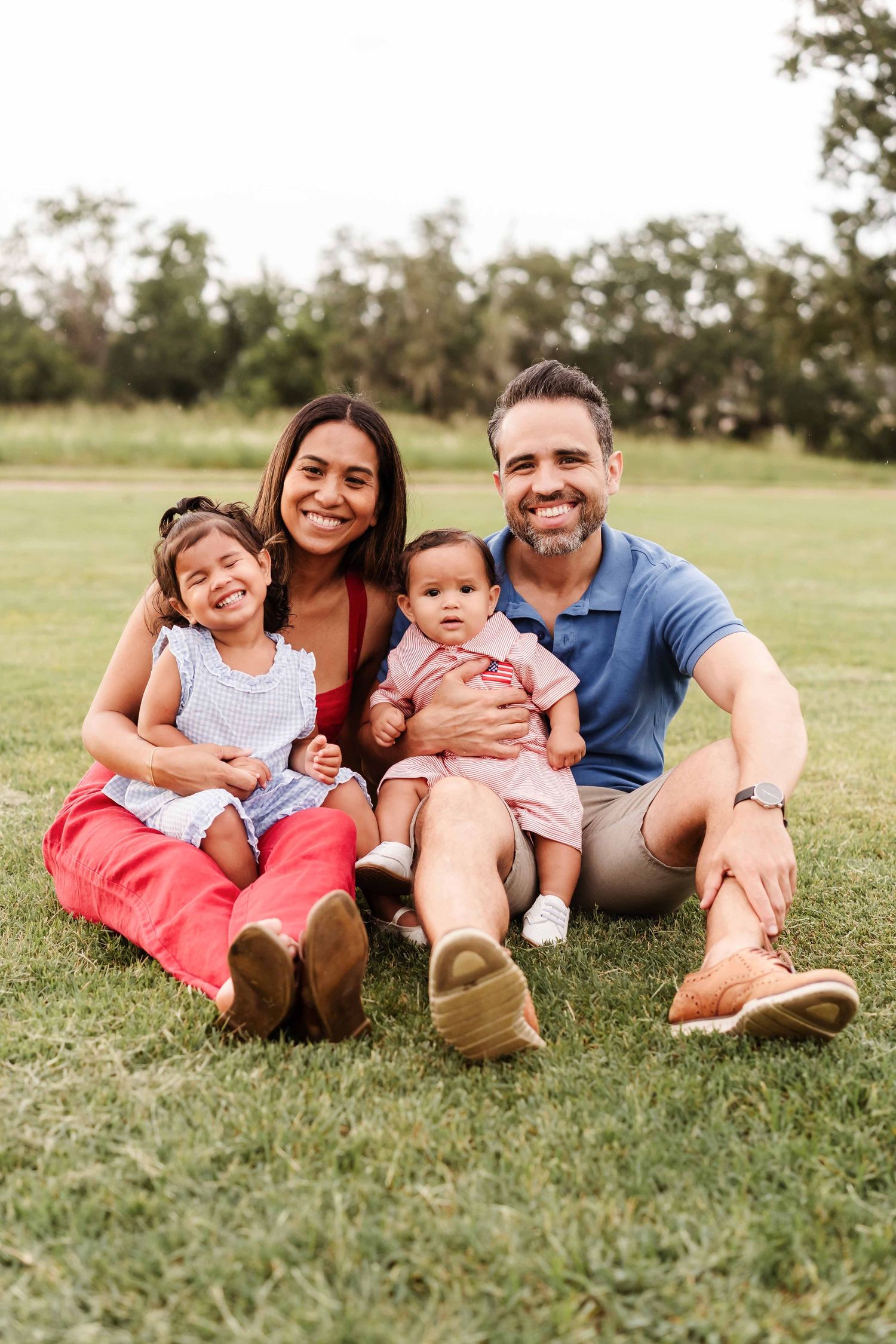 Happy family of four sitting on green grass in a park, parents smiling with two young daughters on a sunny day.