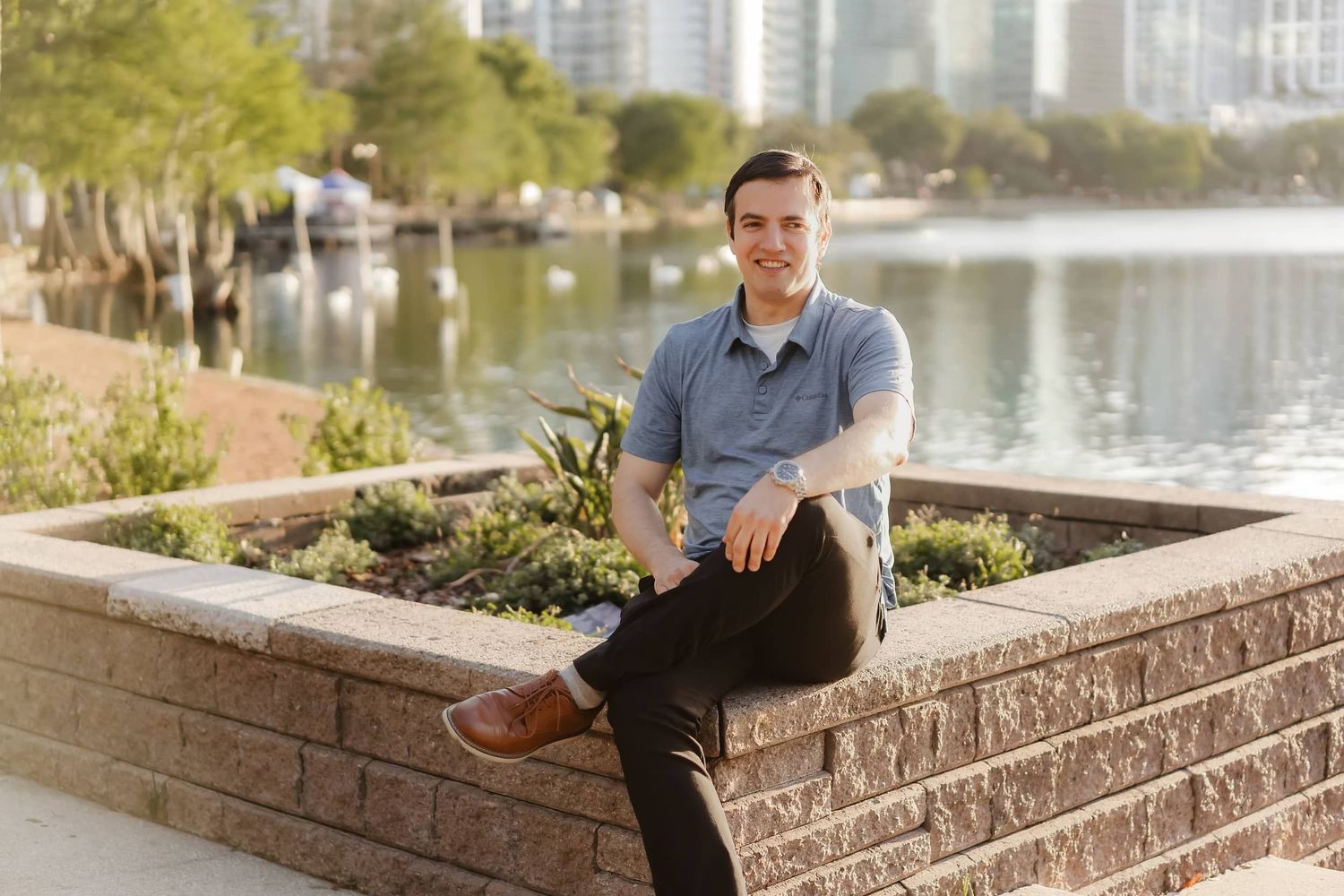 Smiling man sitting on a brick planter outdoors near a waterfront park with city buildings in the background.