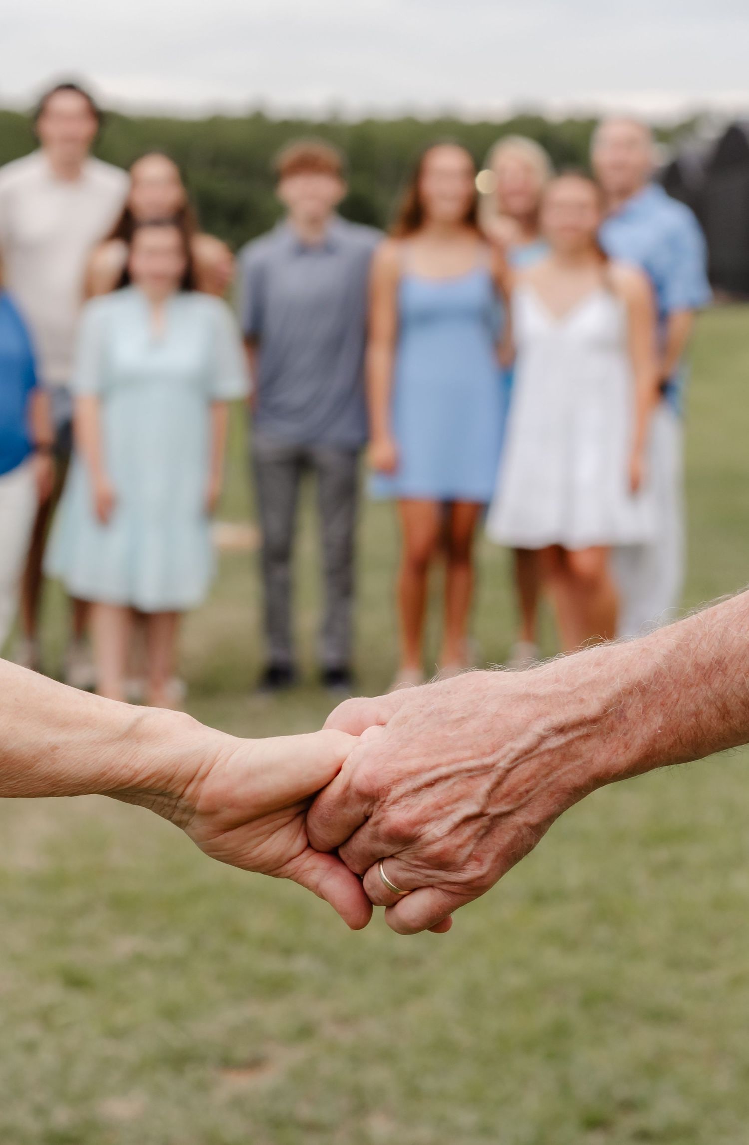 Elderly couple holding hands in foreground with blurred family group standing on green lawn in background.
