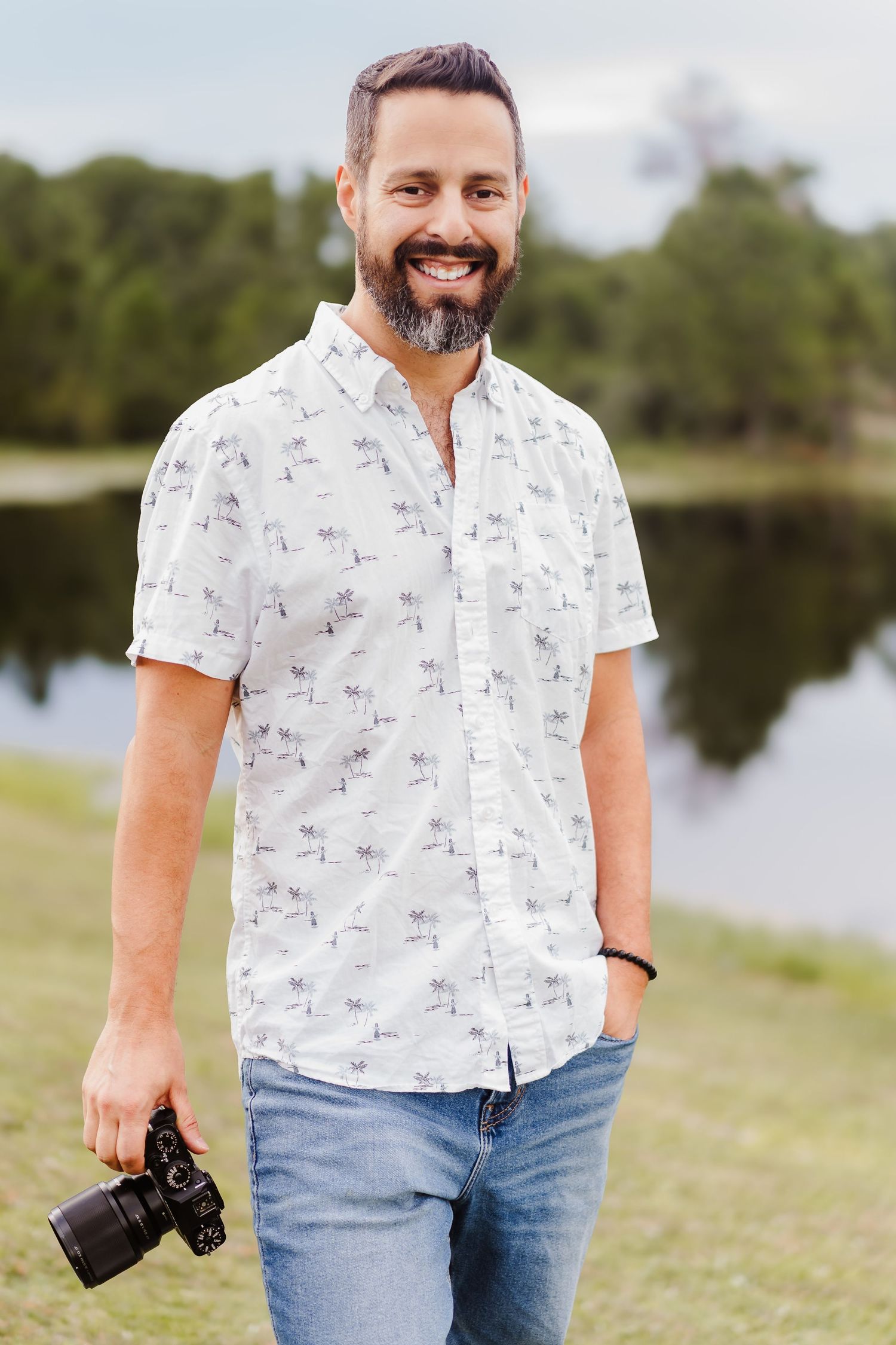 A photographer in a white patterned button-down shirt holds a camera near a scenic lake.