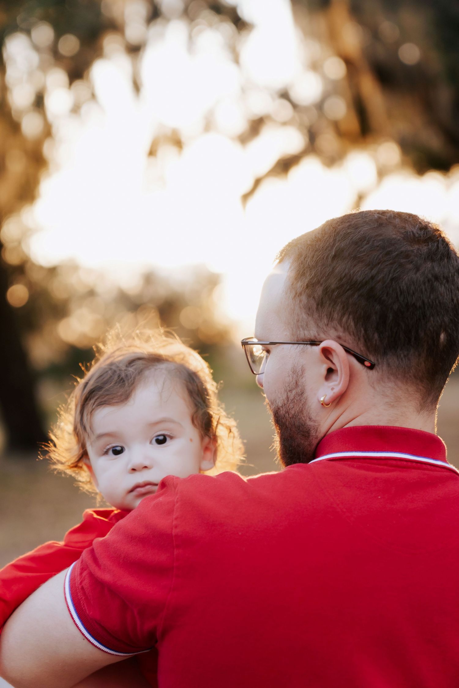 A man in a red polo shirt holds a baby outdoors in warm golden sunlight near trees.